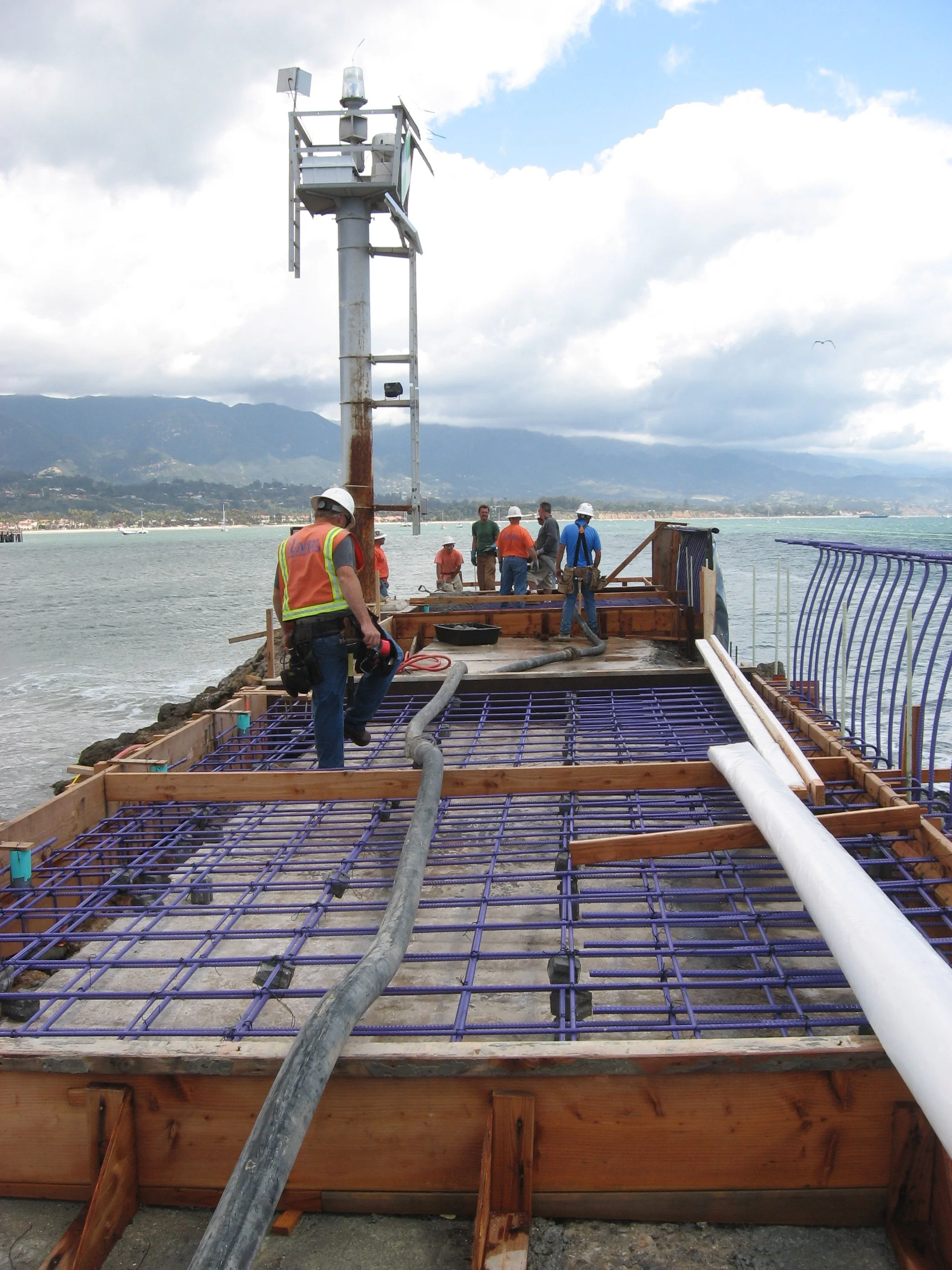 Workers at a construction site by a body of water, installing rebar reinforcement for a concrete structure, with a large drill and a tall metal tower in the background.