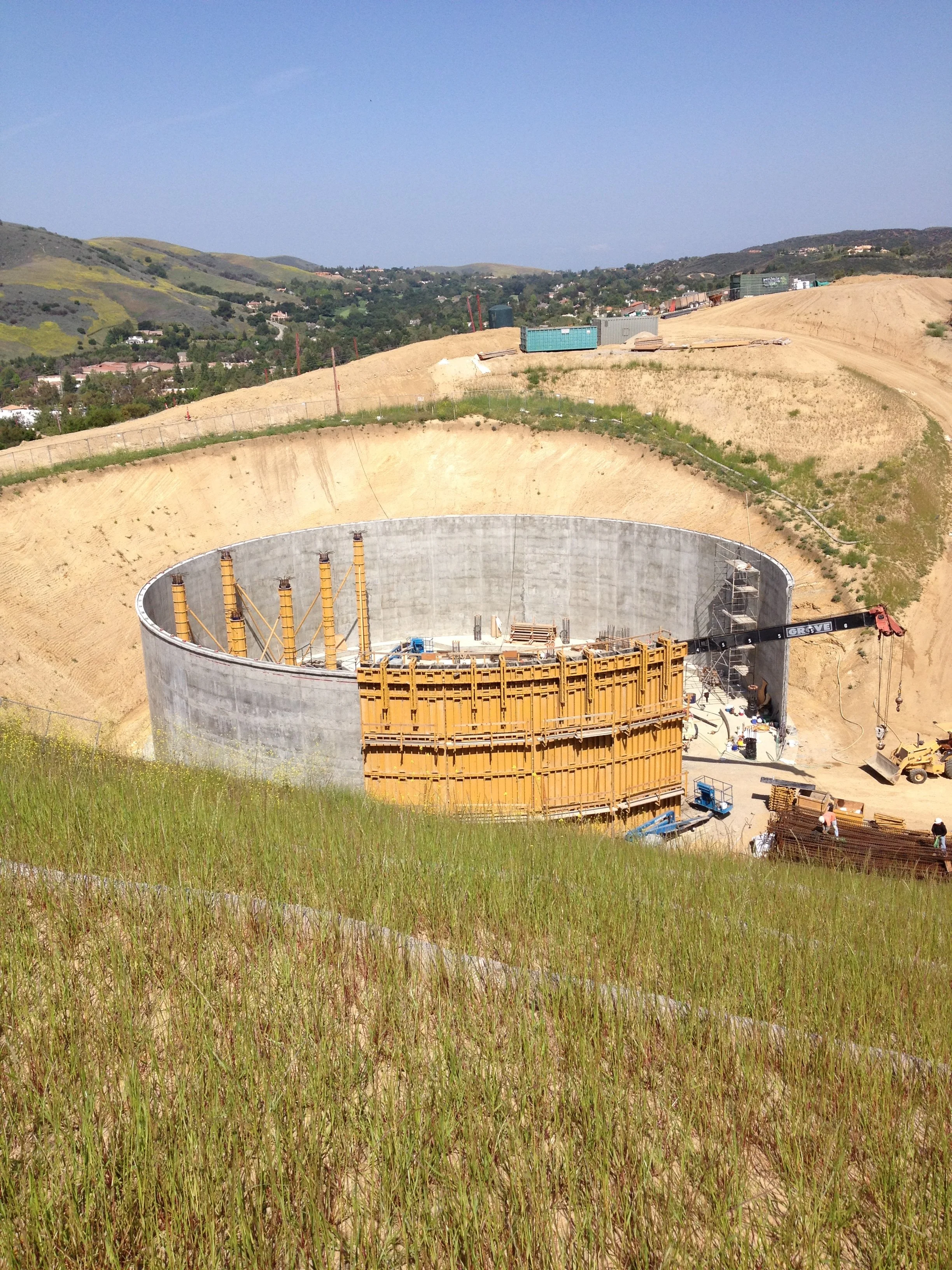 Construction site with a large circular concrete structure being built on a hillside, with construction equipment and materials around.