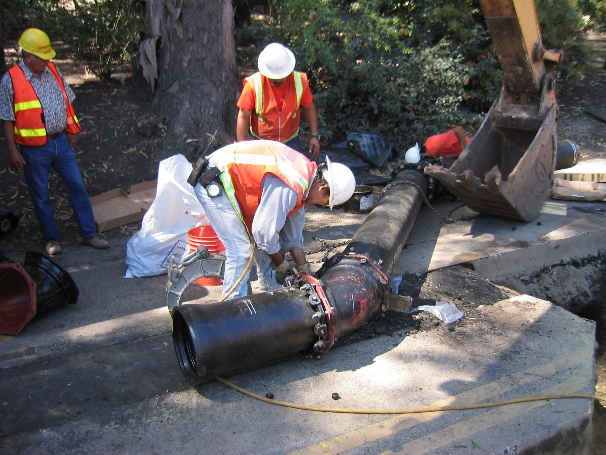 Workers in safety gear, including helmets and reflective vests, assess damage to a large pipe and fallen tree amidst debris after a natural disaster or accident.
