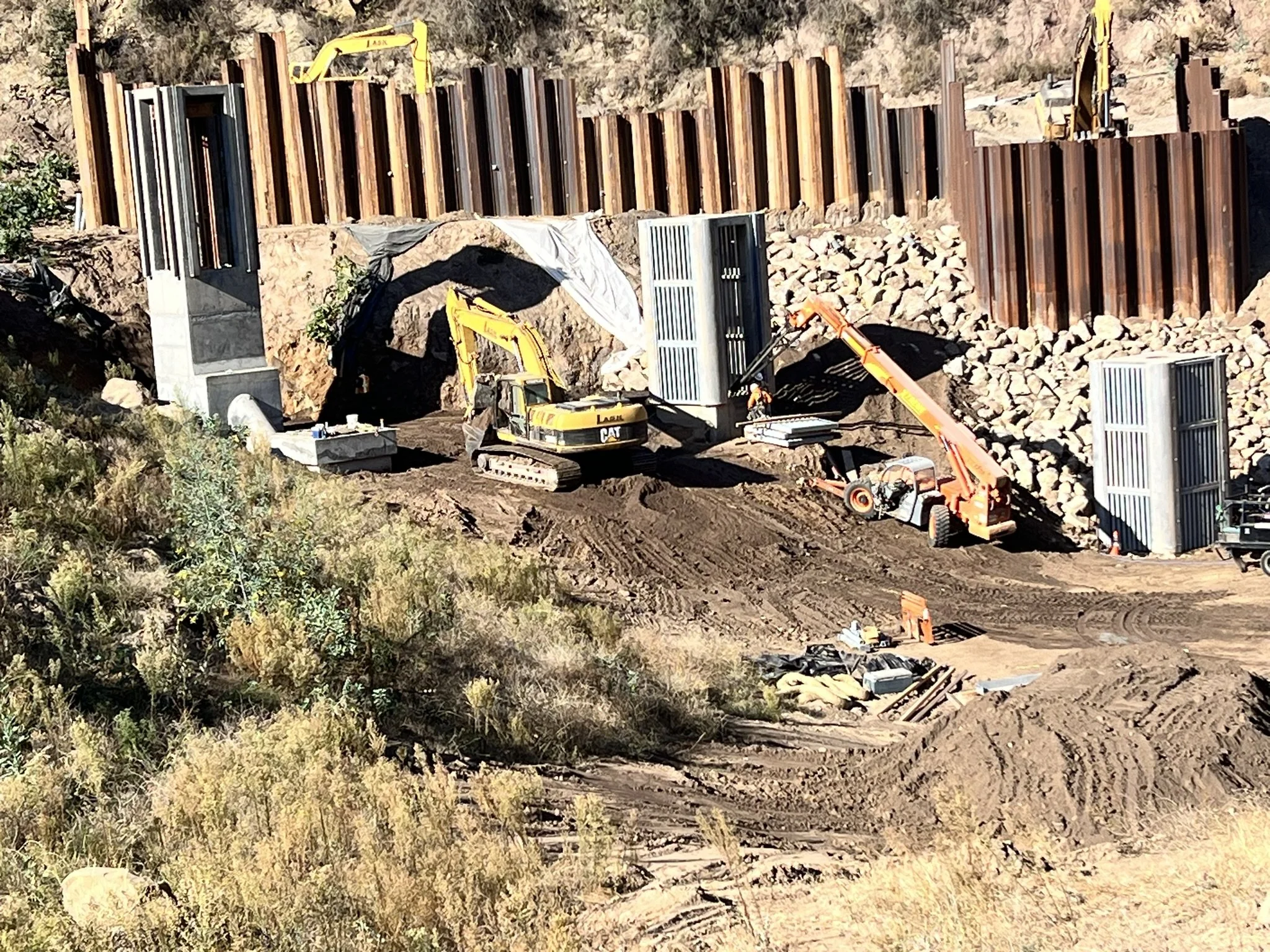 Construction site with a large dirt excavation, steel retaining walls, and yellow construction machinery. Vegetation surrounds the site.
