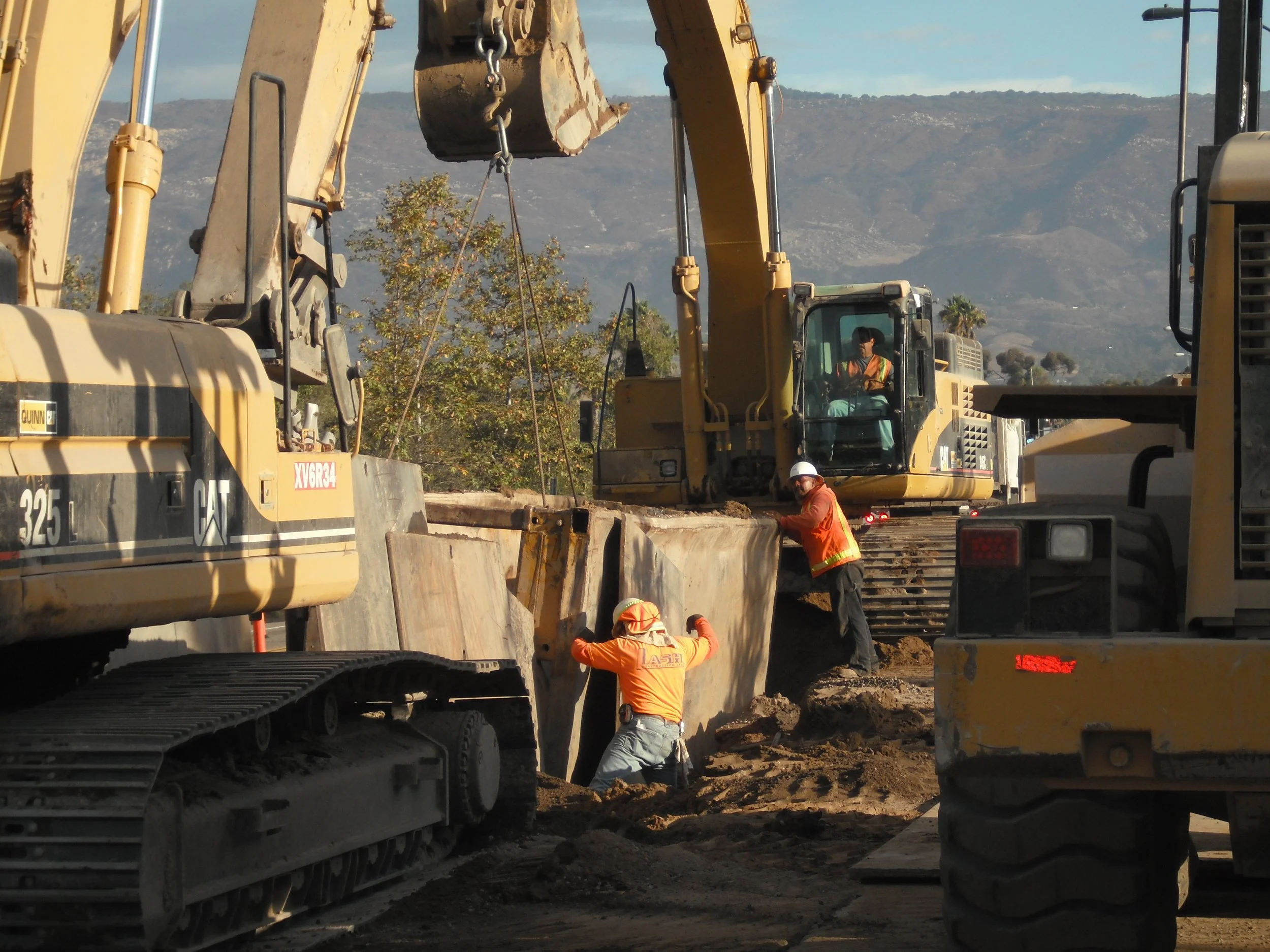 Construction site with multiple yellow excavators and workers wearing safety gear, digging and working on the ground, with hills and trees in the background.