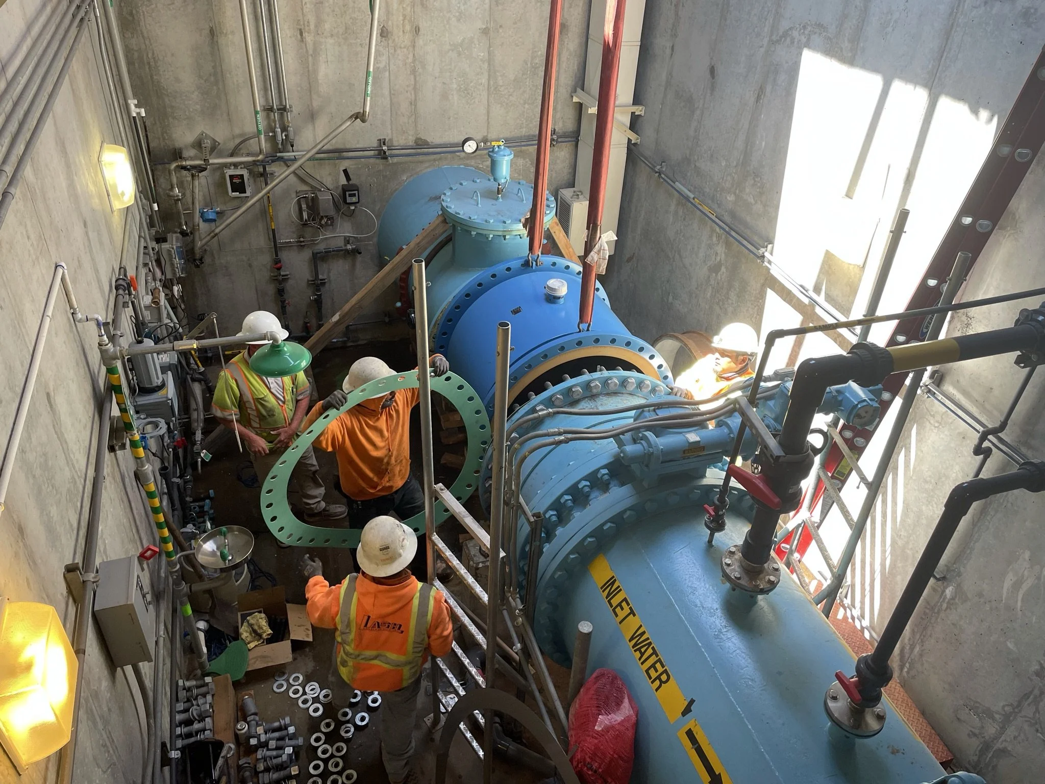 Construction workers installing a large blue pipe in a concrete shaft with scaffolding and various tools and pipes around.