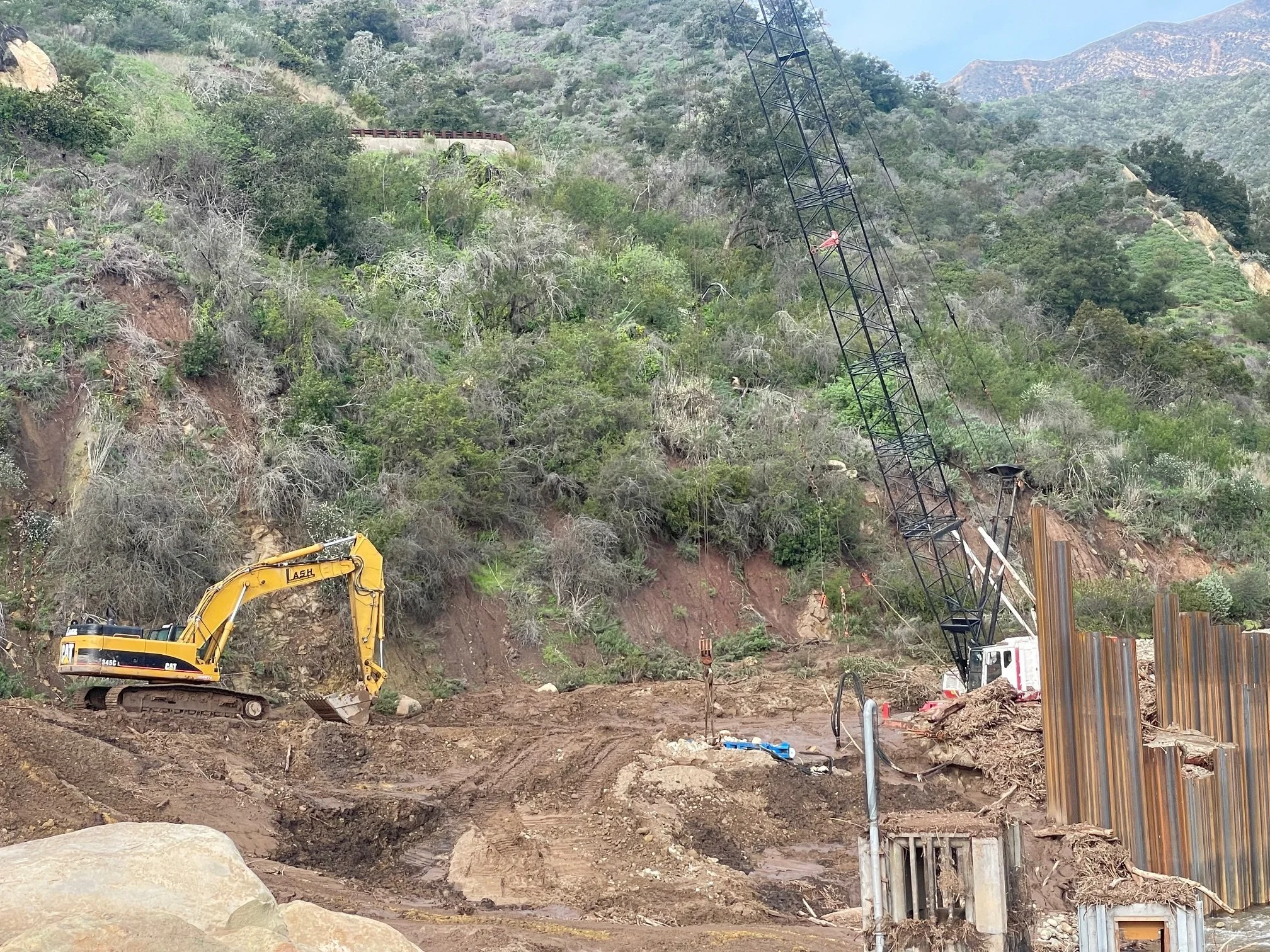 Construction site on a hillside with excavators, a crane, and steel supports, surrounded by trees and mountains.