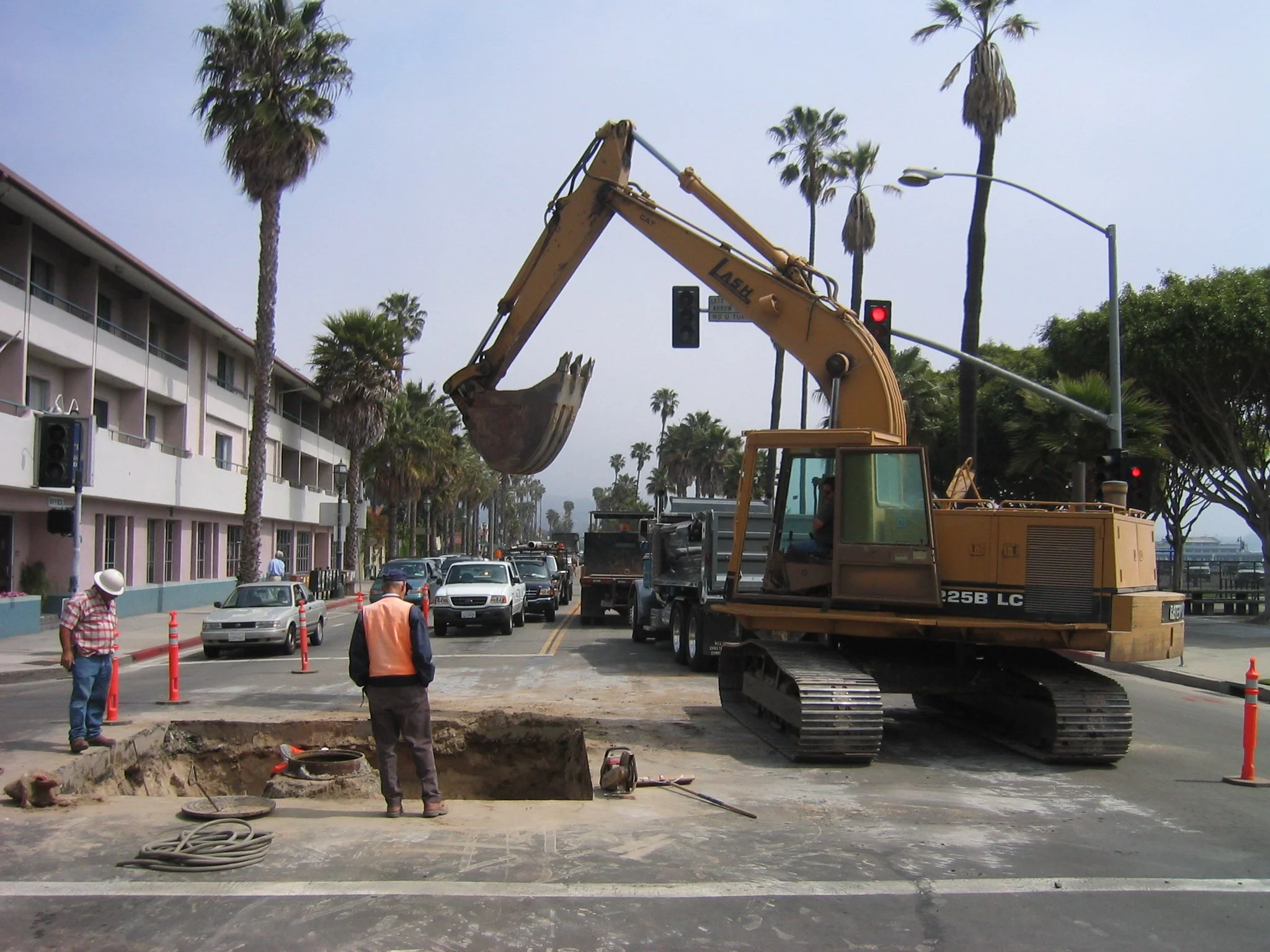 Construction workers working on a road with an excavator digging into the ground, blocking part of the street, during daytime with palm trees and buildings in the background.