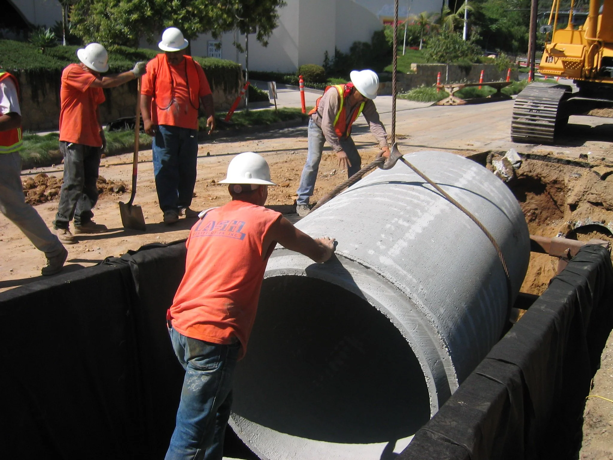 Construction workers wearing orange shirts and white helmets installing large concrete pipe underground at a construction site with an excavator in the background.