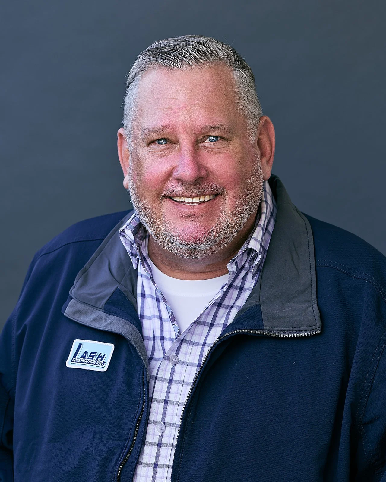 Portrait of a smiling middle-aged man with a beard and gray hair, wearing a navy blue jacket over a plaid shirt, against a solid gray background.