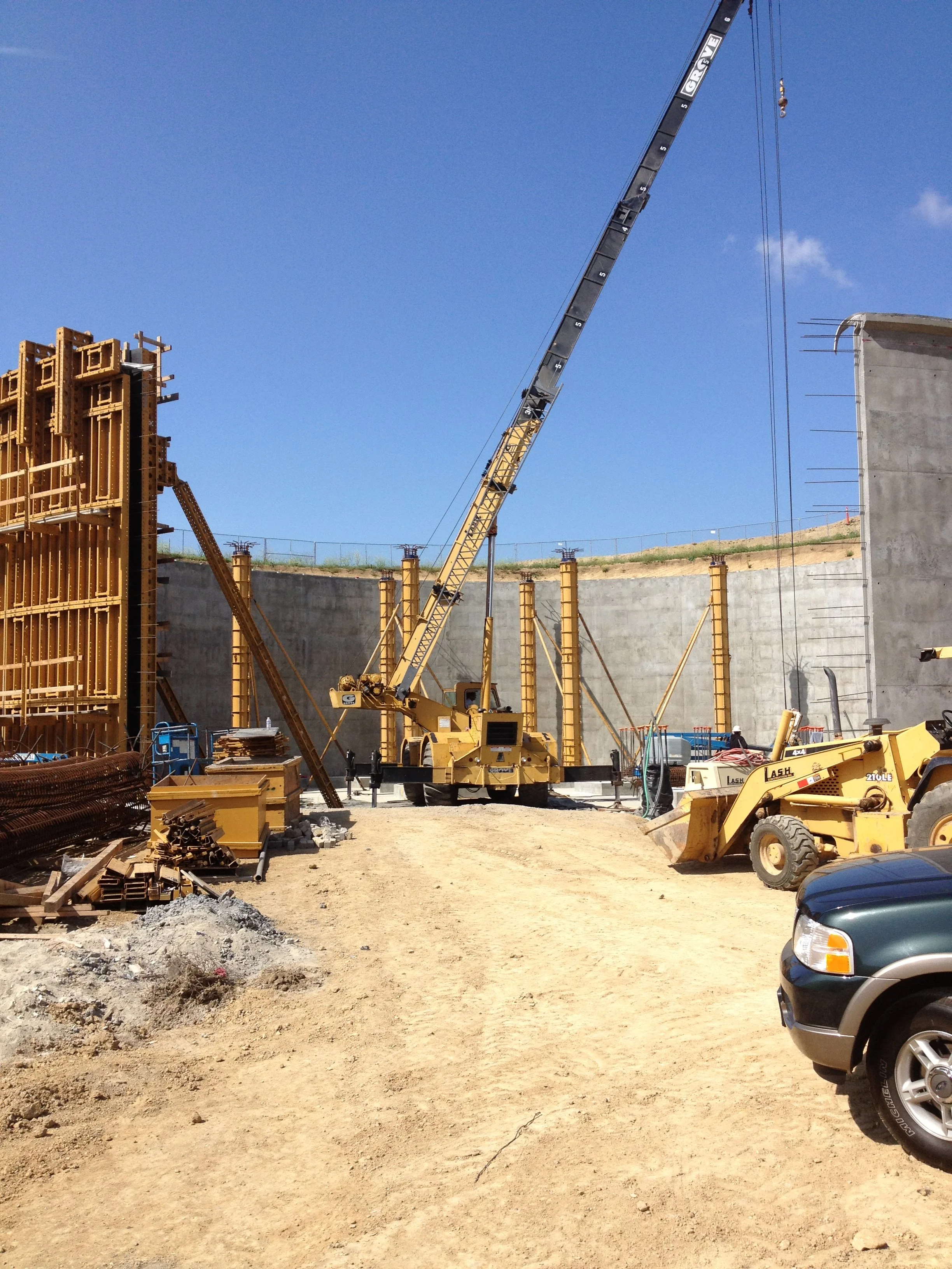 View of a construction site with a tall crane, construction materials, and a partially built concrete wall under a clear blue sky.