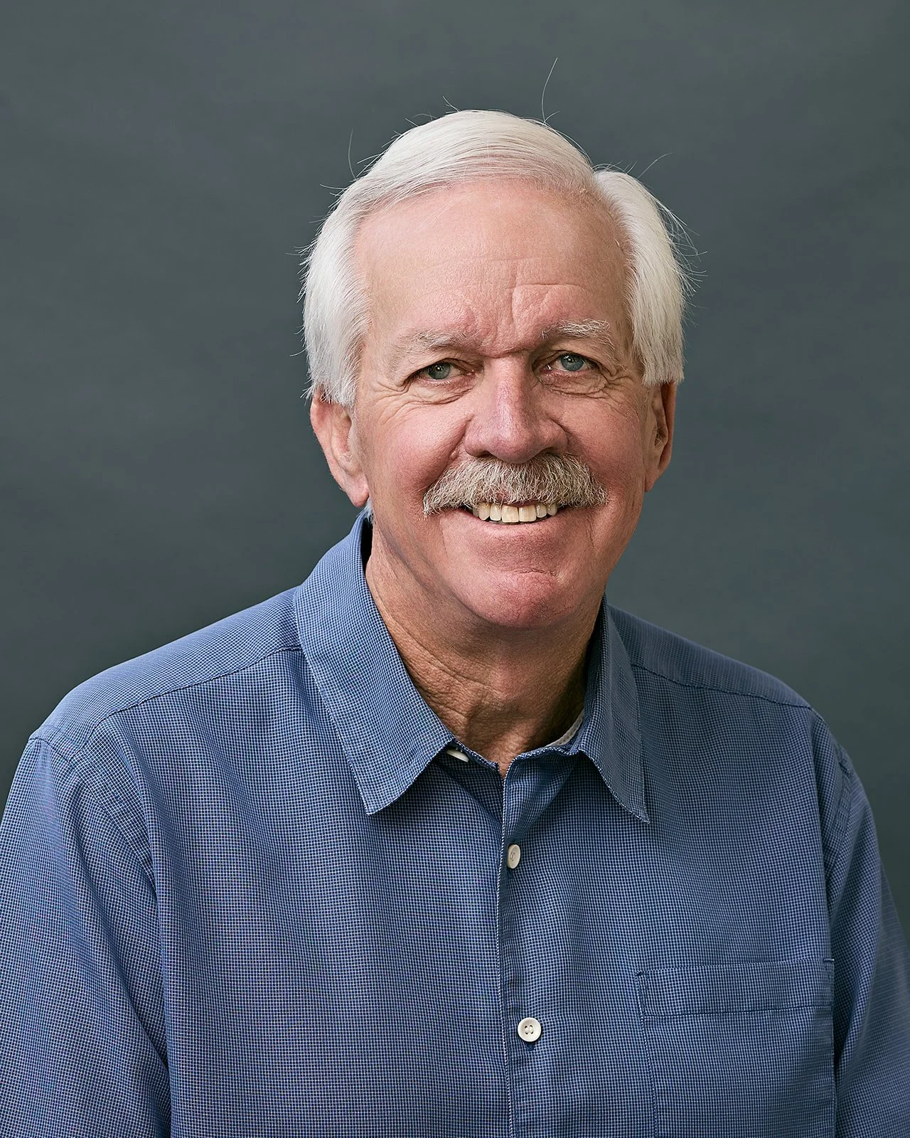 A portrait of an older man with white hair and a mustache, smiling, wearing a blue checkered shirt, against a dark gray background.