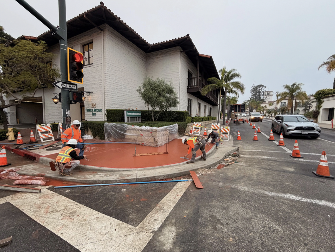 Construction workers applying a fresh layer of red asphalt on a section of sidewalk near a building and busy street, with traffic cones and barriers for safety.