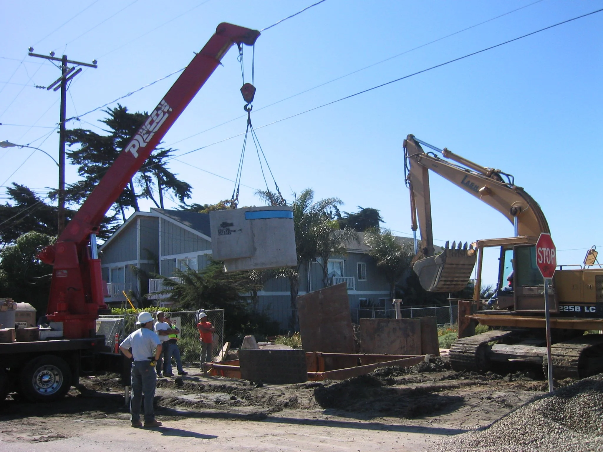 Construction workers and heavy machinery working on a construction site, lifting a large concrete block into place near houses with trees and power lines in the background.