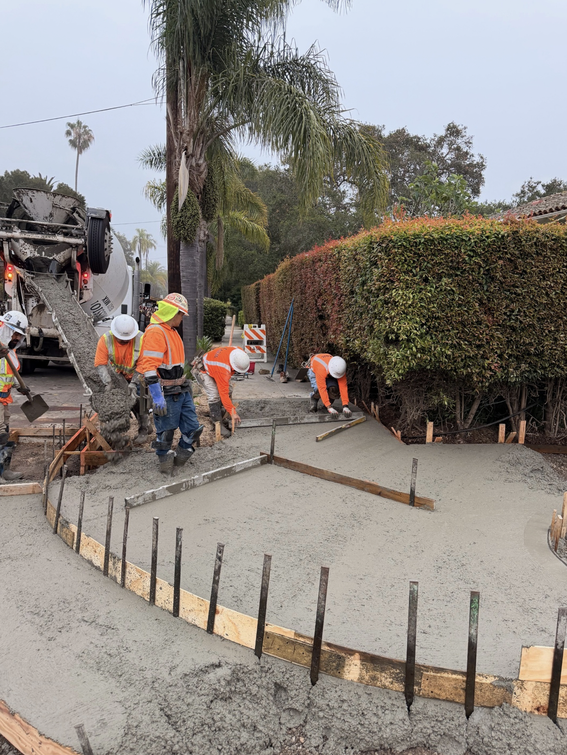 Construction workers in orange safety vests and white helmets pouring and leveling concrete for a sidewalk, with a truck pouring concrete nearby, in a residential area with trees and bushes.