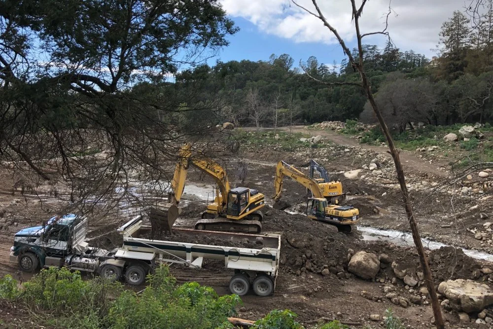 Construction site with two yellow excavators and a dump truck working on a muddy, rocky landscape surrounded by trees.