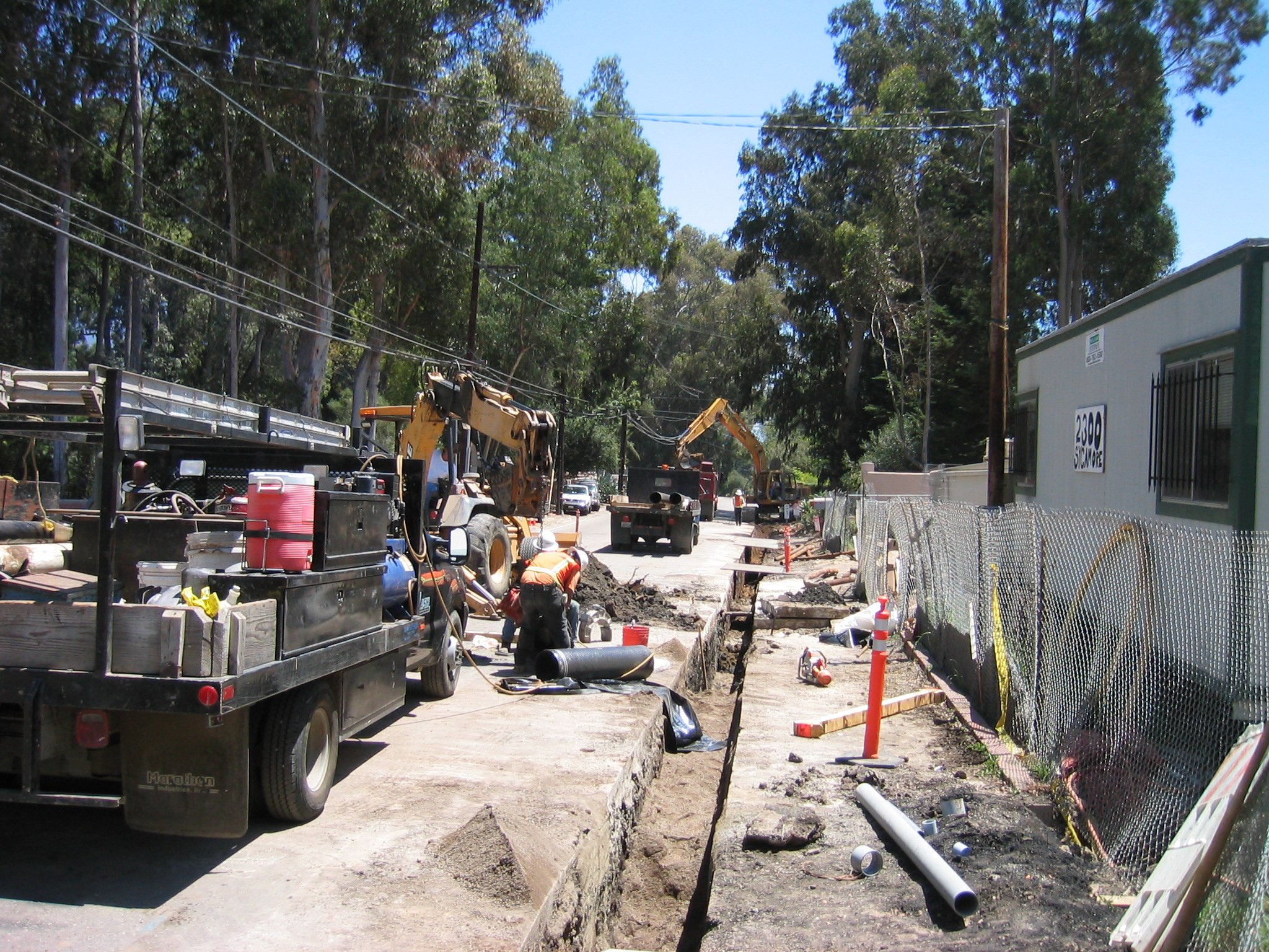 Construction workers and equipment working on a street repair project, with utility poles and trees in the background.