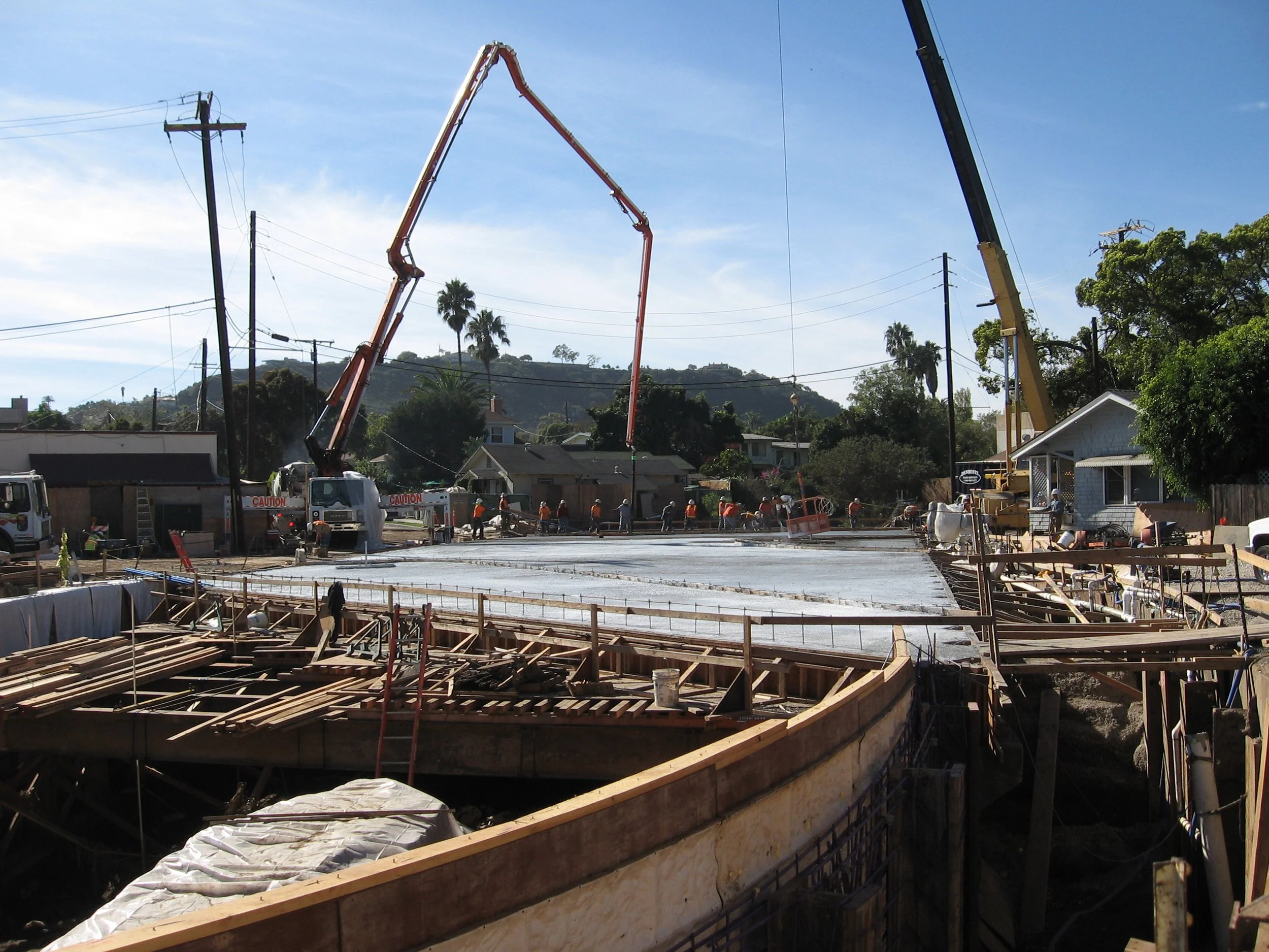 Construction site with workers, concrete, and cranes installing foundation in a residential area with houses and trees in the background.