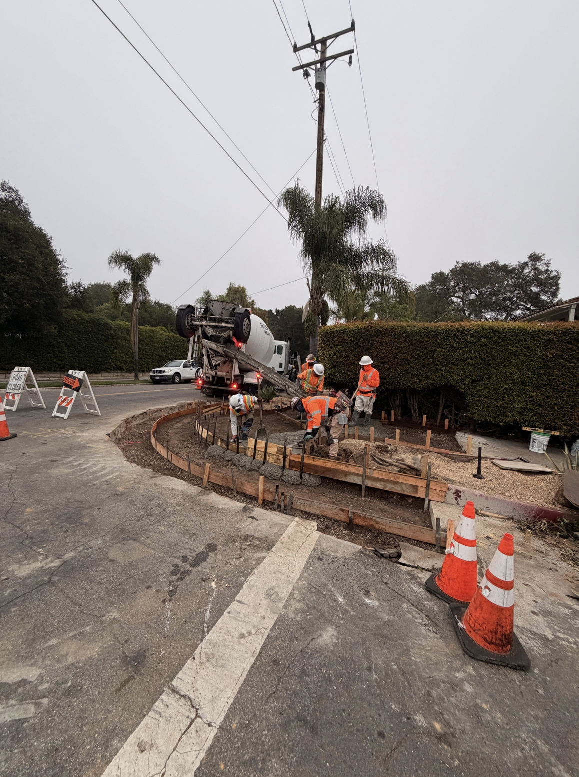 Construction workers wearing safety gear are working on a road repair project, using a concrete mixer truck and wooden forms to pour new concrete around a curved section of the street. Traffic cones and signs are placed to block off the area.