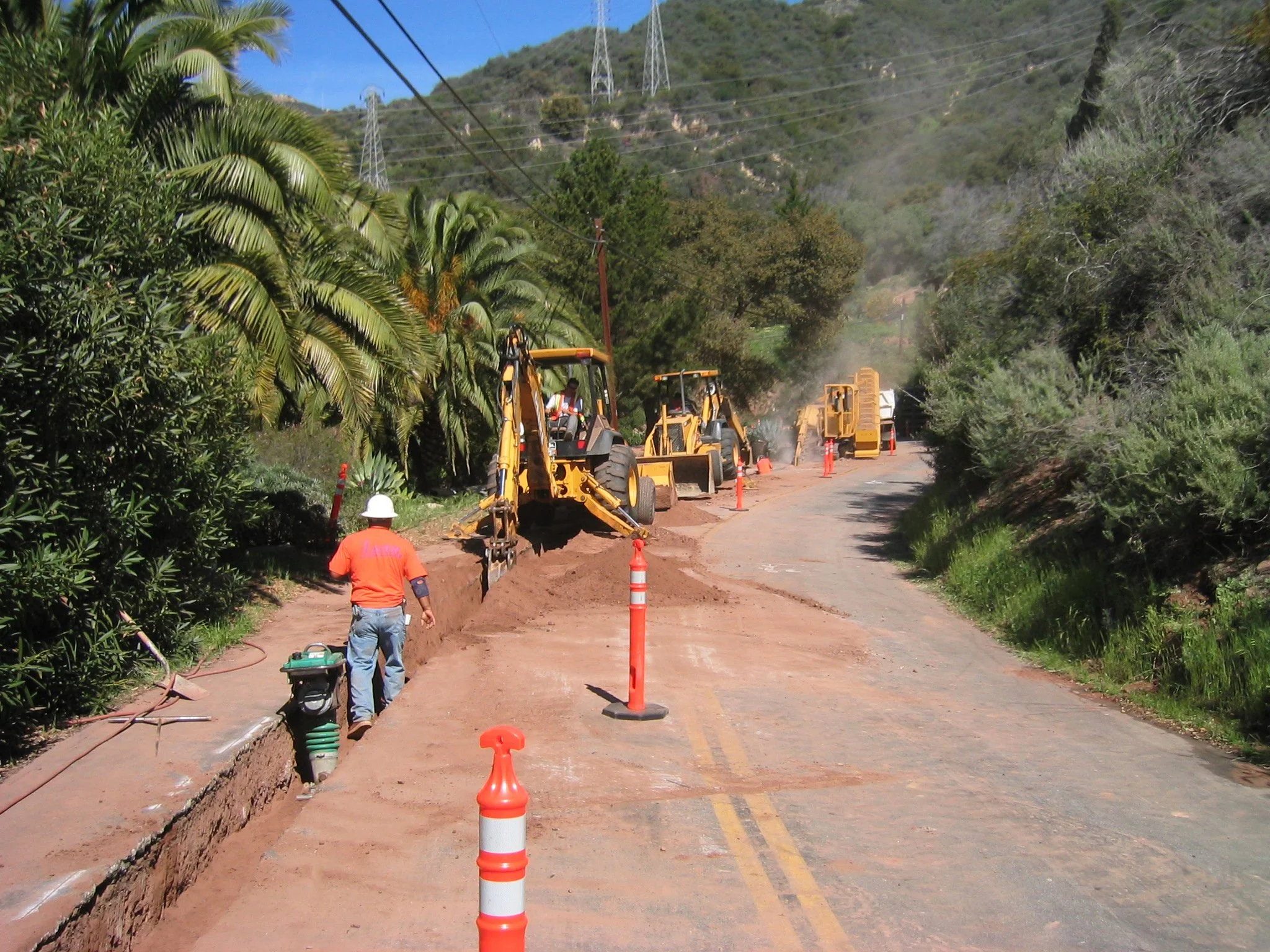 Road construction with orange traffic cones, workers operating machinery, and dirt being worked on, with trees on either side of the road and hills in the background.