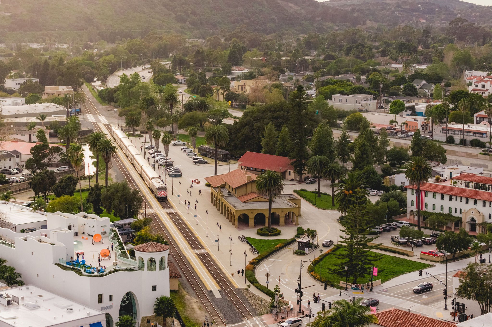 Aerial view of a suburban area with a train station, train tracks, and surrounding houses and buildings. There are trees and greenery throughout the area.