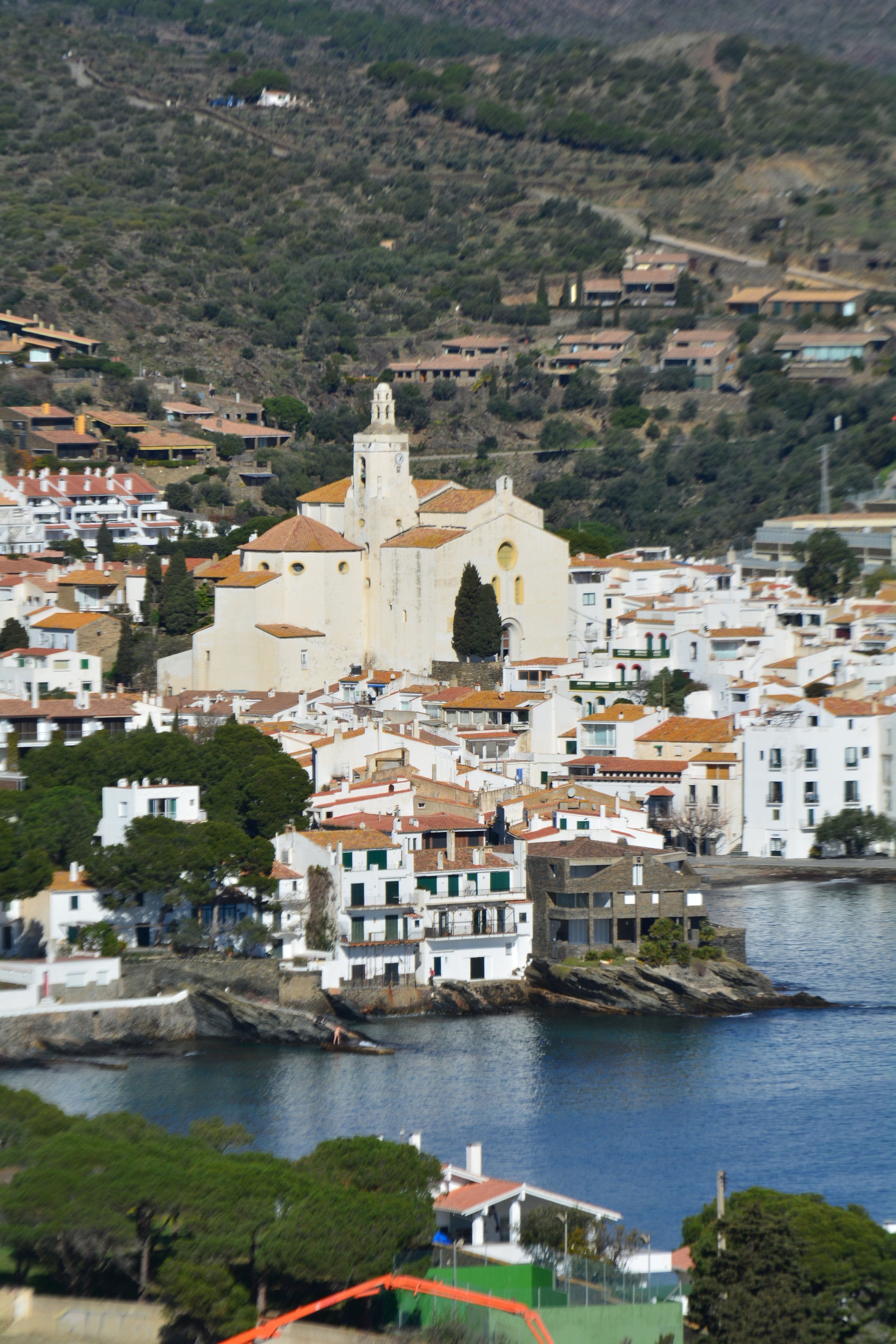 Vista panorámica de un pueblo costero con una iglesia blanca en el centro, rodeada de casas blancas con techos rojos y vegetación, en una zona montañosa con un cuerpo de agua en primer plano.