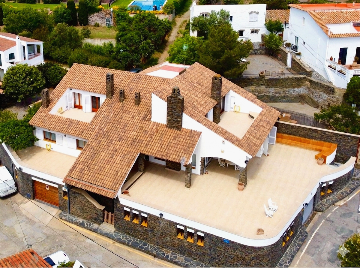 Casa grande con techo de tejas de color marrón, paredes blancas y chimeneas de ladrillo. Amplio patio con sillas y ventanas. Rodeada de árboles y otras casas blancas.