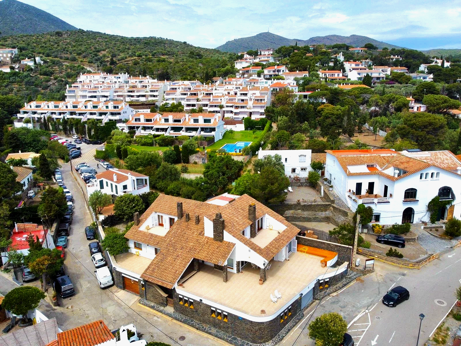 Vista aérea de un barrio residencial en una zona montañosa con casas blancas, un área con piscina y muchas casas con techos de teja, rodeadas de árboles y vegetación.
