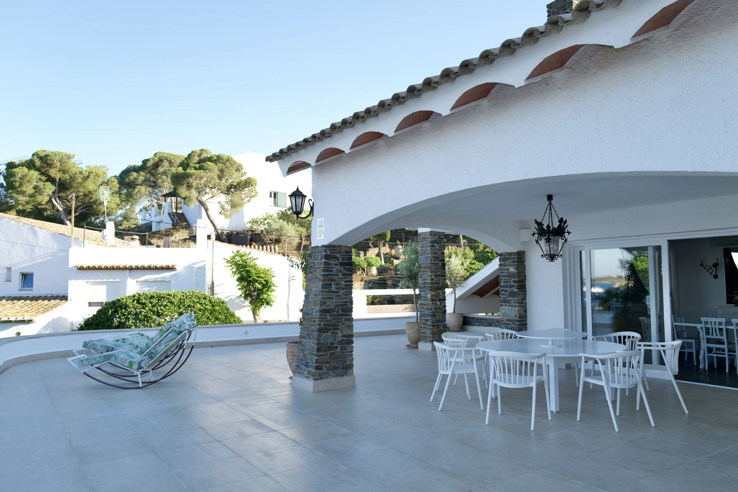 Terraza de casa con mesas y sillas blancas, lámpara de hierro decorativa, columnas con piedra, y fondo de árboles y casas blancas bajo cielo despejado.