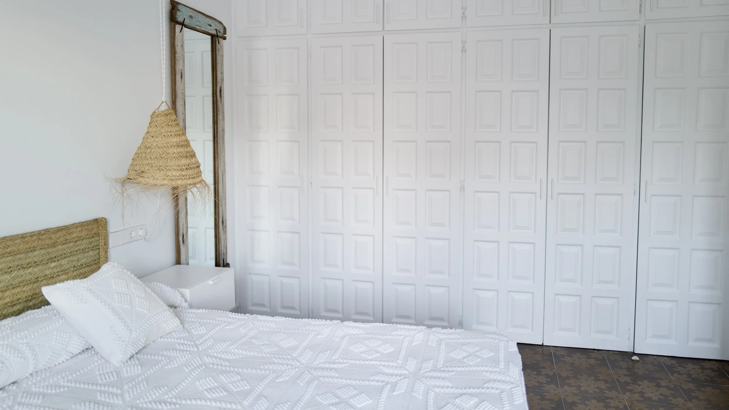Cuarto con pared de armario empotrado blanco y cama con cabecera de mimbre y sábanas blancas, colgador de mimbre y espejo de madera en la pared.