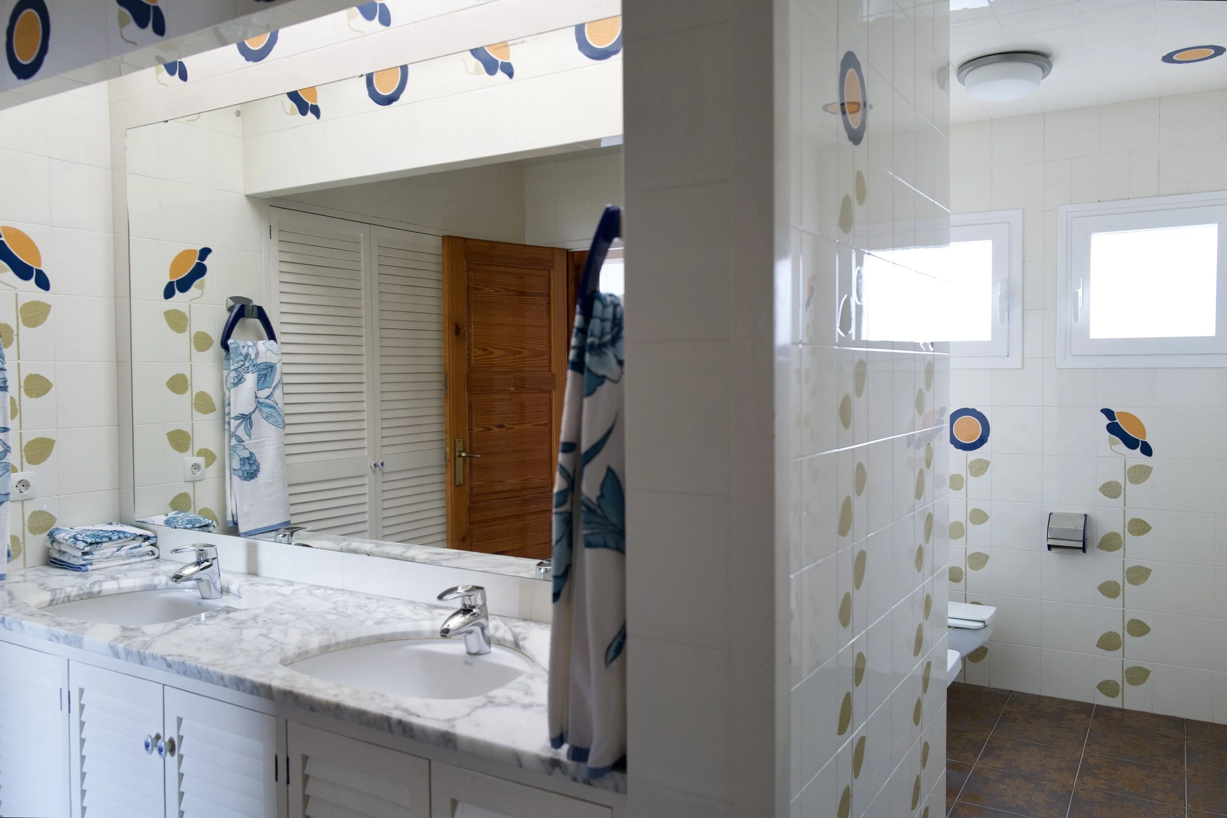 Un baño con doble lavabo de mármol, un espejo, toallas blancas con azul, lavadero de ducha con cortina y paredes decoradas con galletas azules, naranjas y verdes.