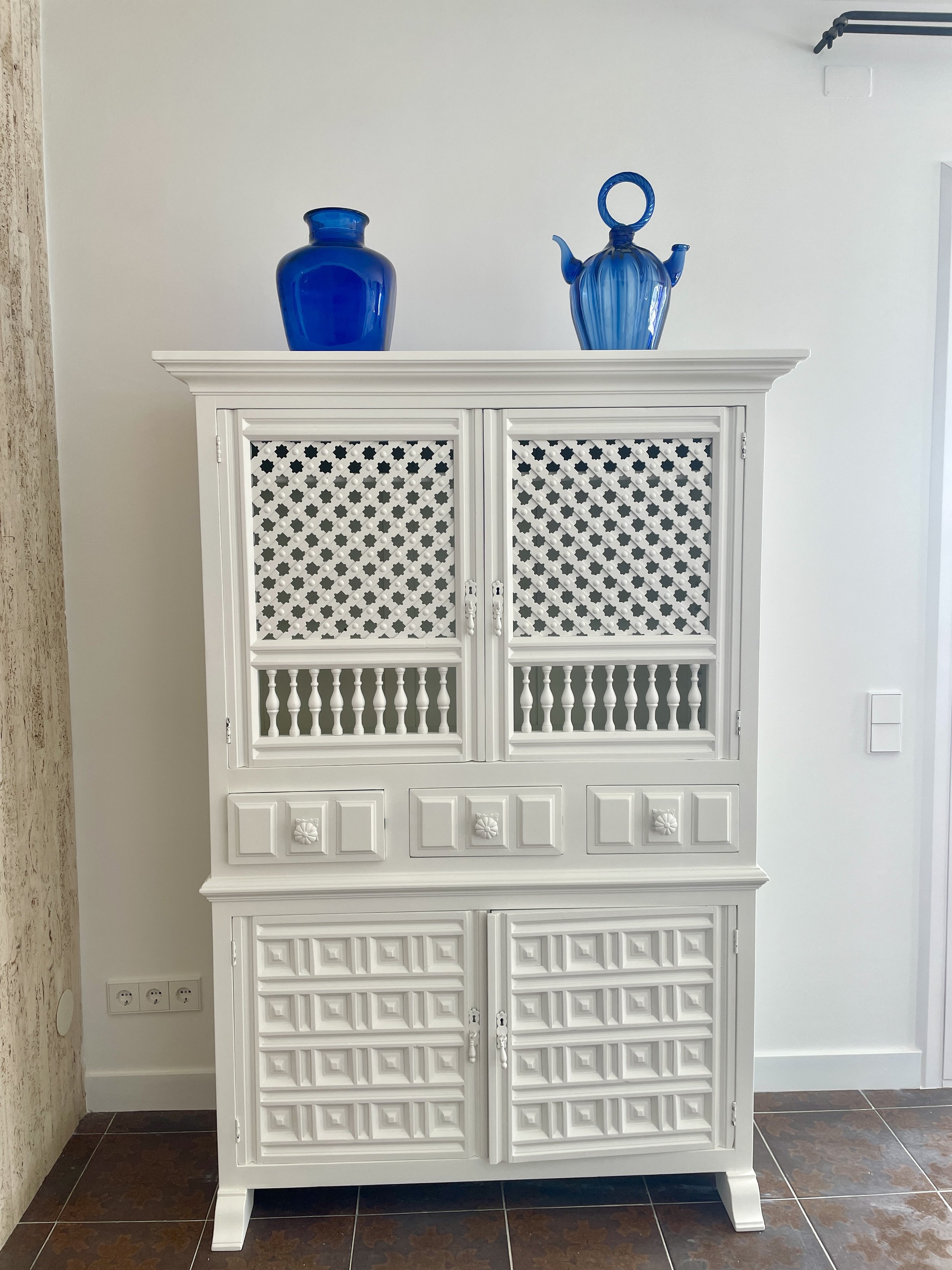 Estantería blanca con puertas de rejilla y decoración, en la parte superior hay dos frascos de vidrio azul, uno sólido y otro con diseño rayado y asa