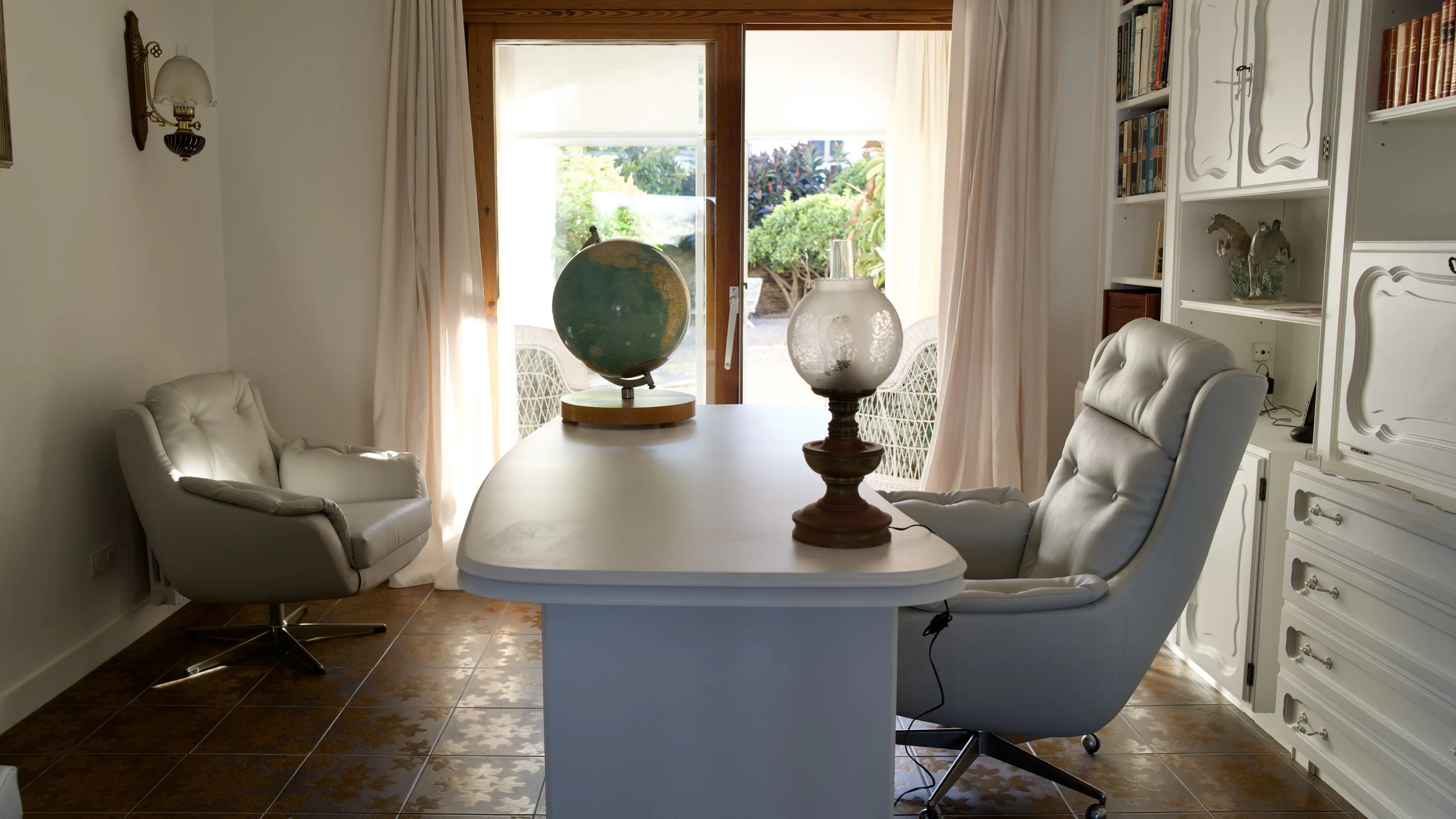Sala de estar luminosa con mesa ovalada blanca, dos sillones blancos tapizados, una lámpara grande y un globo terráqueo sobre la mesa, puertas corredizas con cortinas y un fondo con plantas verdes afuera.