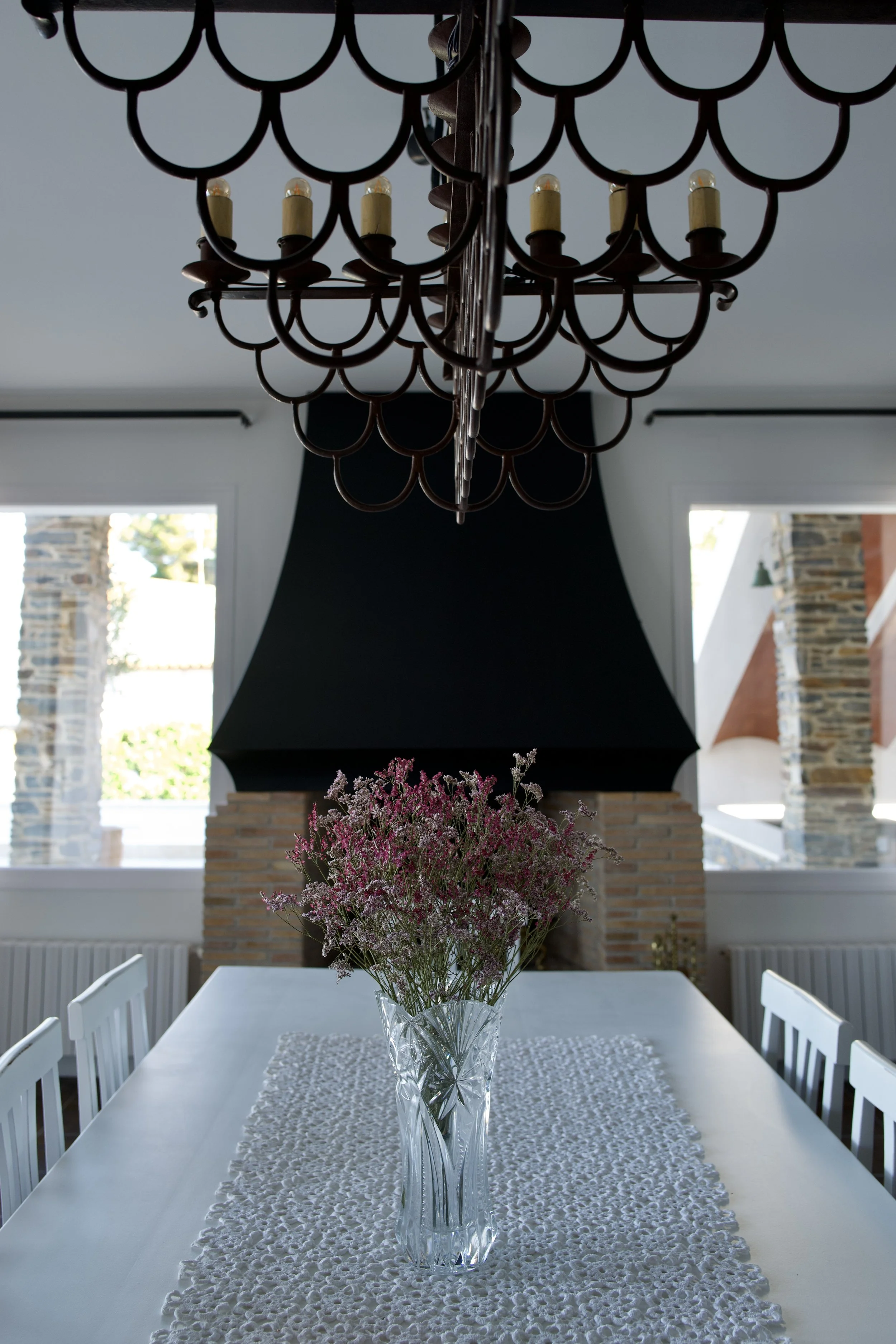 Mesa blanca con un jarrón de cristal con flores rosadas y moradas, en un comedor con ventana y chimenea de ladrillo.