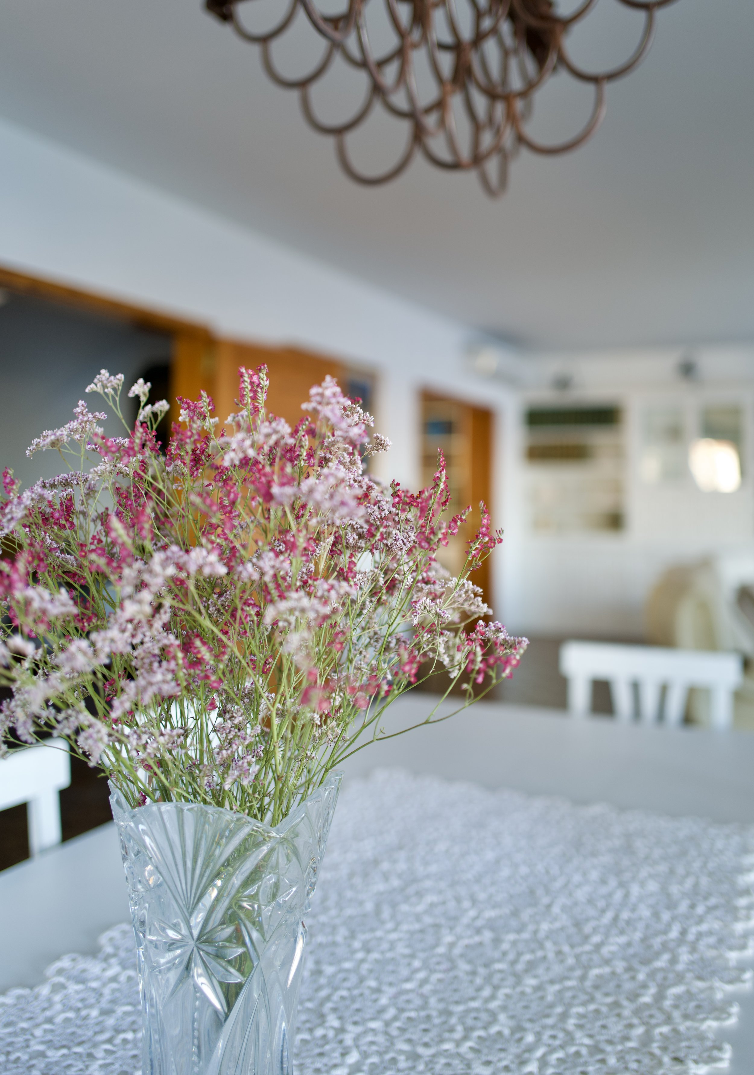 Ramo de flores rosadas y blancas en un jarrón de cristal sobre una mesa cubierta con un mantel de encaje, en un comedor con paredes blancas y ventanas.