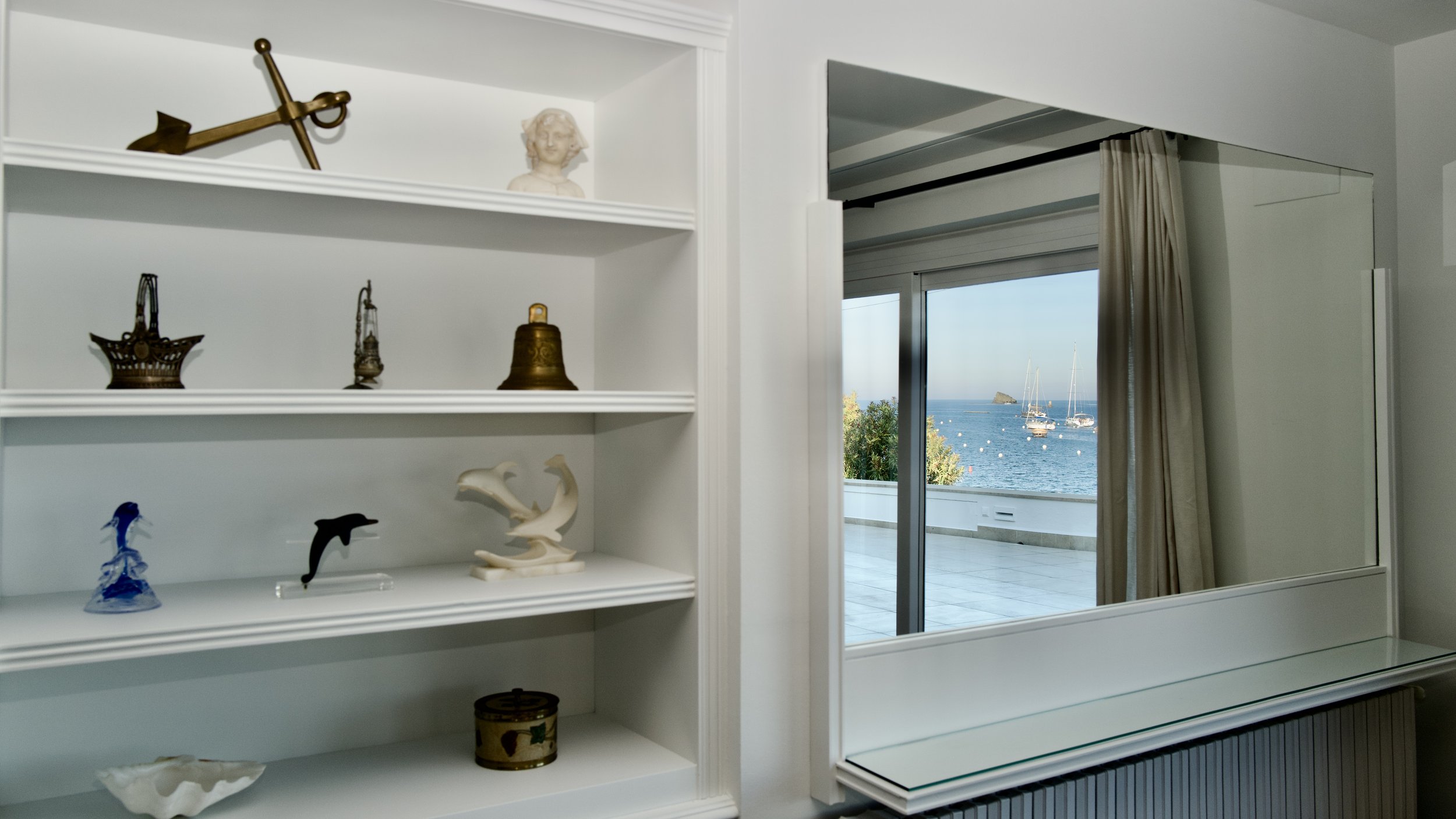 Estanterias y reflejo del mar de la badía de Cadaqués en la Sala de estar de Ca la Nuri Cadaqués, una casa de alquiler de ensueño.