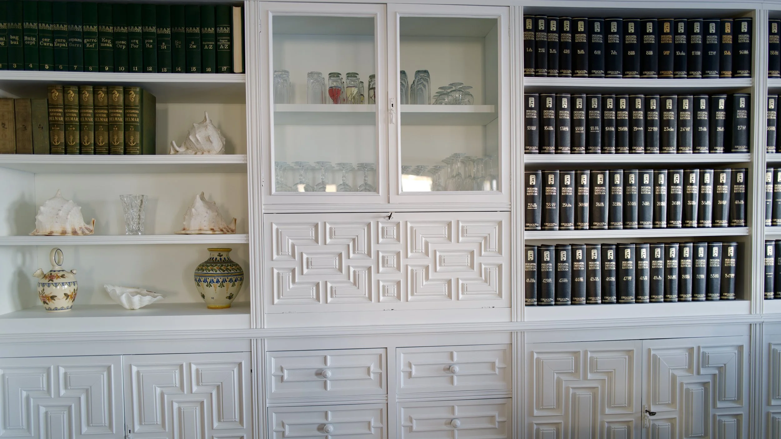 Estantería blanca con libros latinográficos y objetos decorativos como conchas, jarrones y vasos de vidrio.