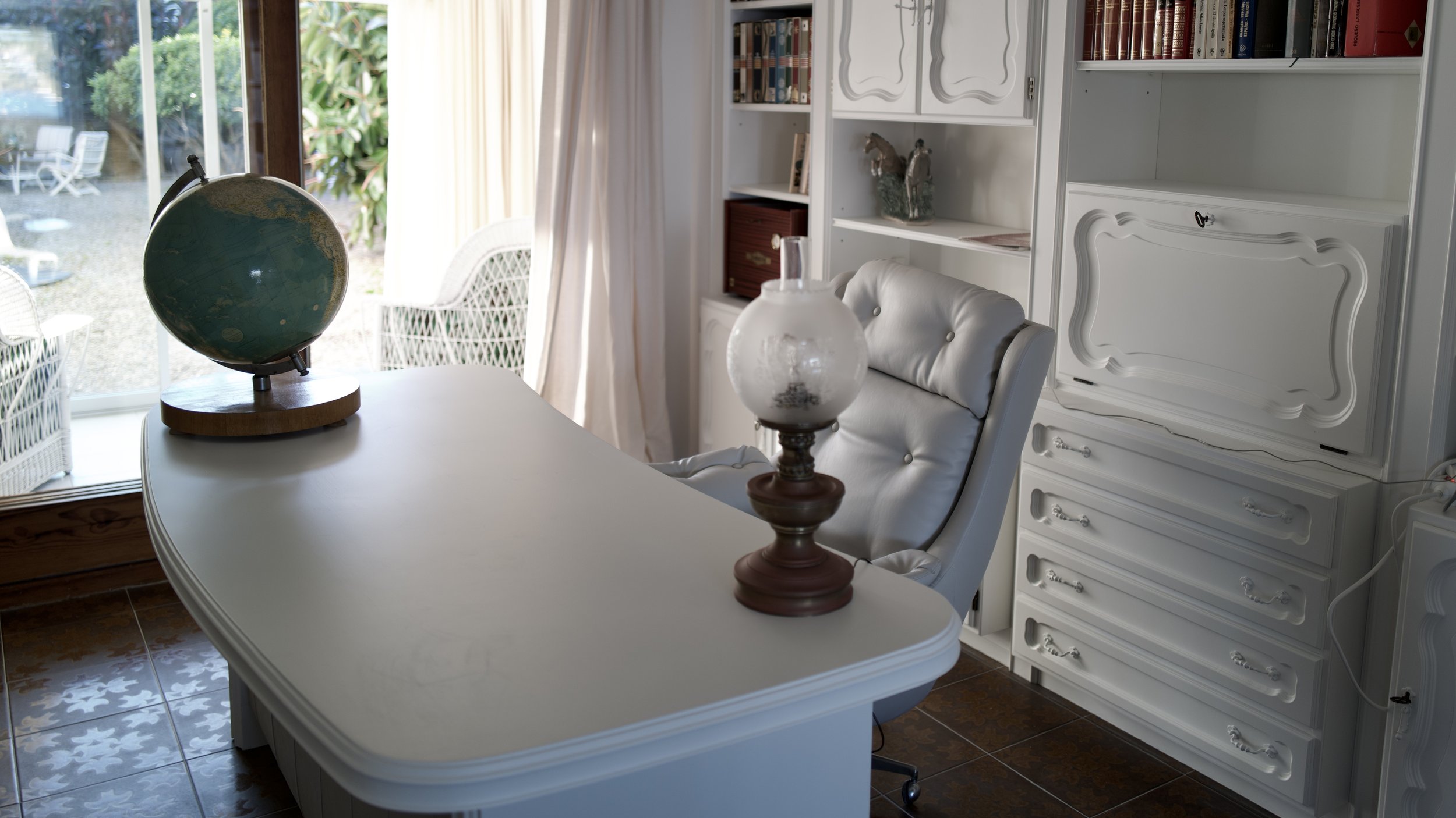Una sala con mesa blanca, una lámpara de vidrio sobre la mesa, un sillón blanco, una estantería blanca con libros y decoraciones, y una ventana con cortinas que da a un jardín exterior con sillas blancas.