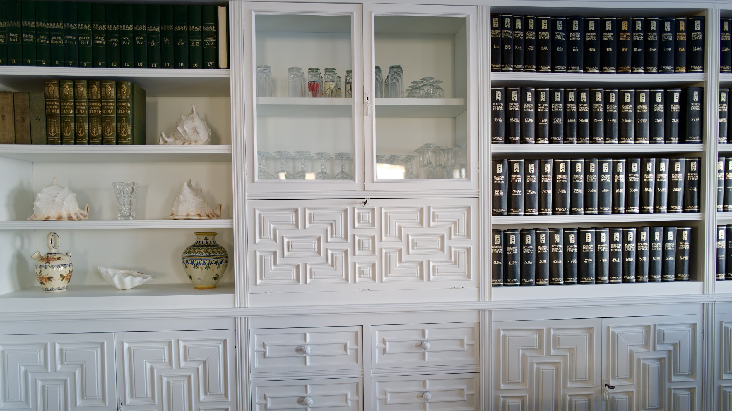 Estantería blanca con libros, cristalería y objetos decorativos en Ca la Nuri, una casa de alquiler con una ubicación única en Cadaqués.