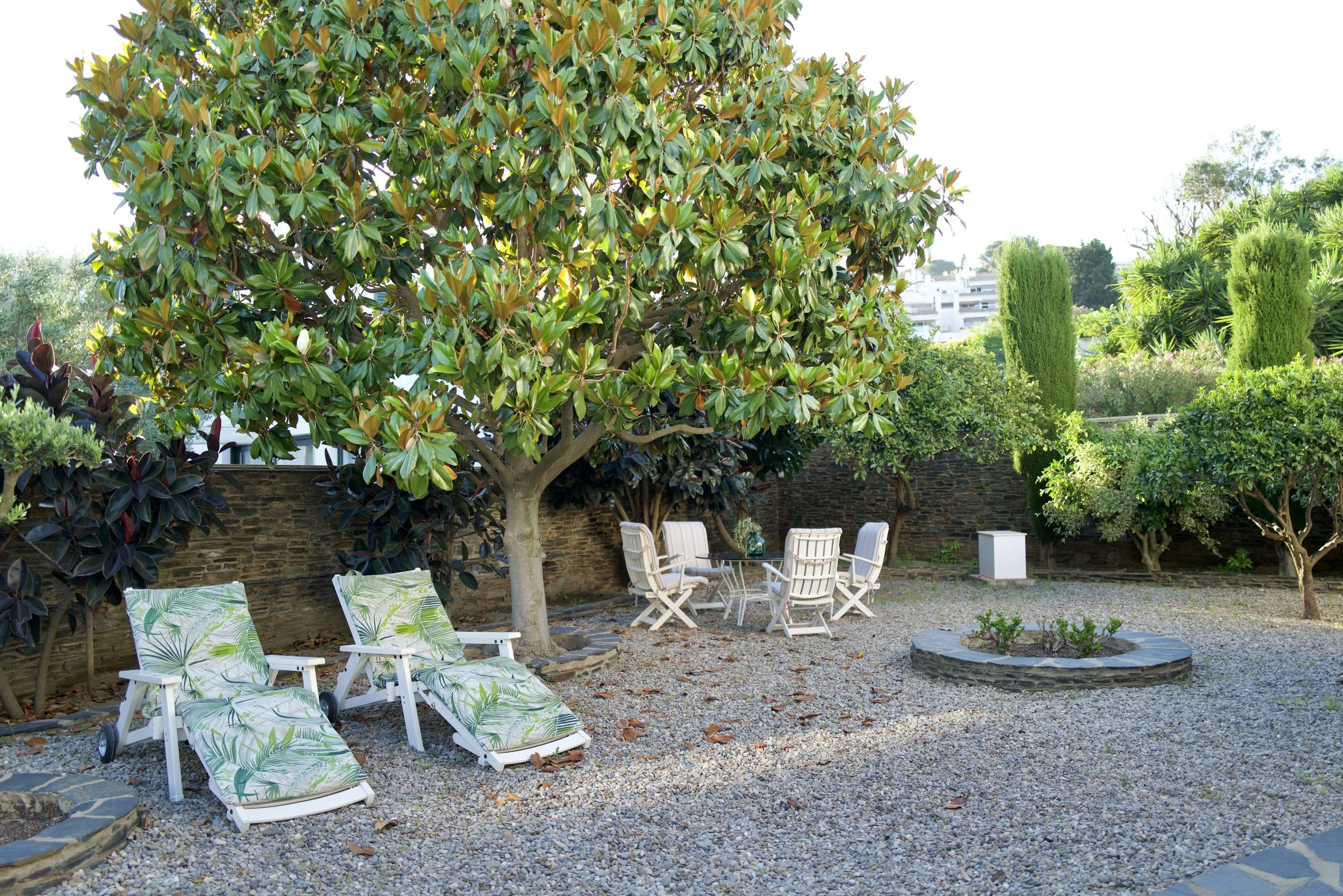 Jardín con varias sillas y mesas de exterior junto a un árbol grande y arbustos verdes, con edificios en el fondo.
