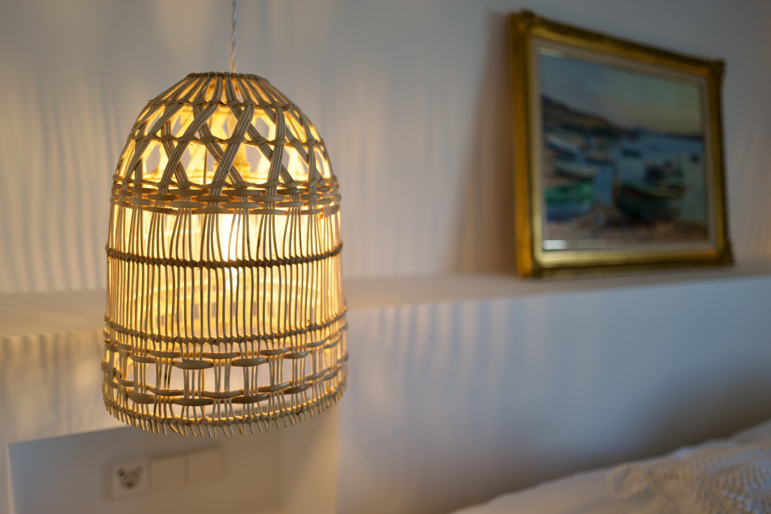 Lámpara de mimbre en forma de cúpula que emite luz cálida, colgada del techo en una habitación, con un cuadro enmarcado en la pared de fondo.