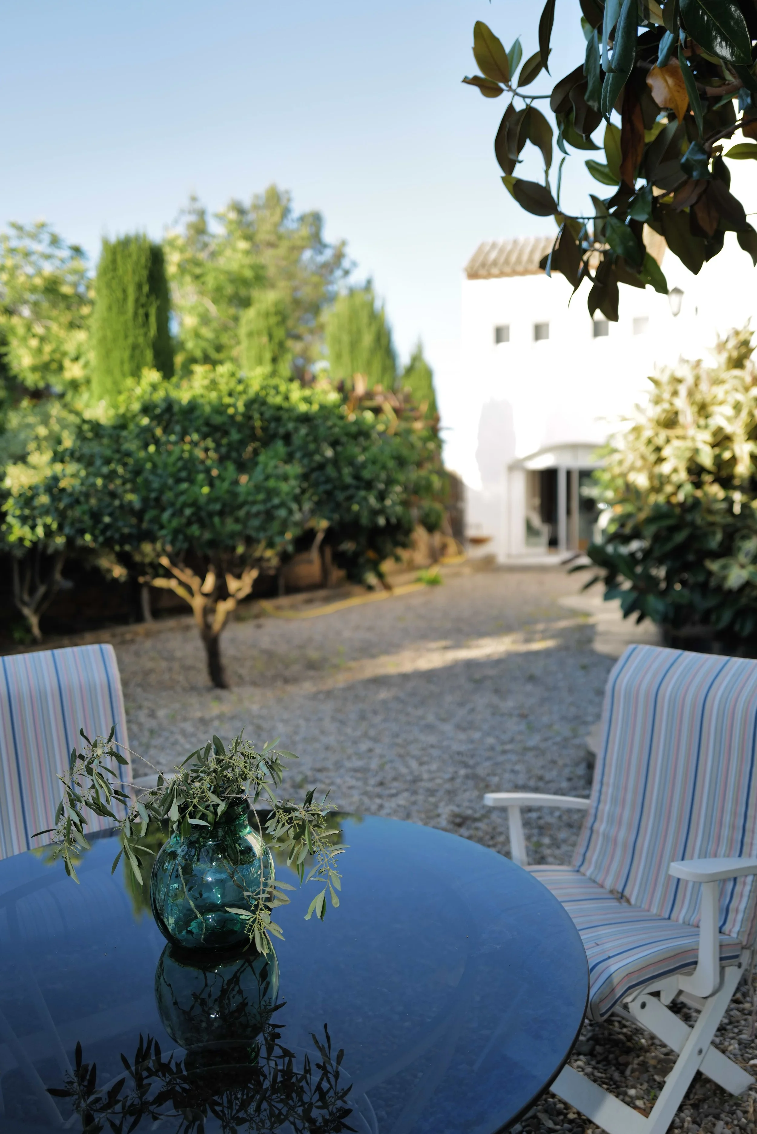 Número de mesa azul con un florero de vidrio con ramas y hojas verdes, en un patio con sillas de plástico con asientos a rayas, árboles y una casa blanca en el fondo.