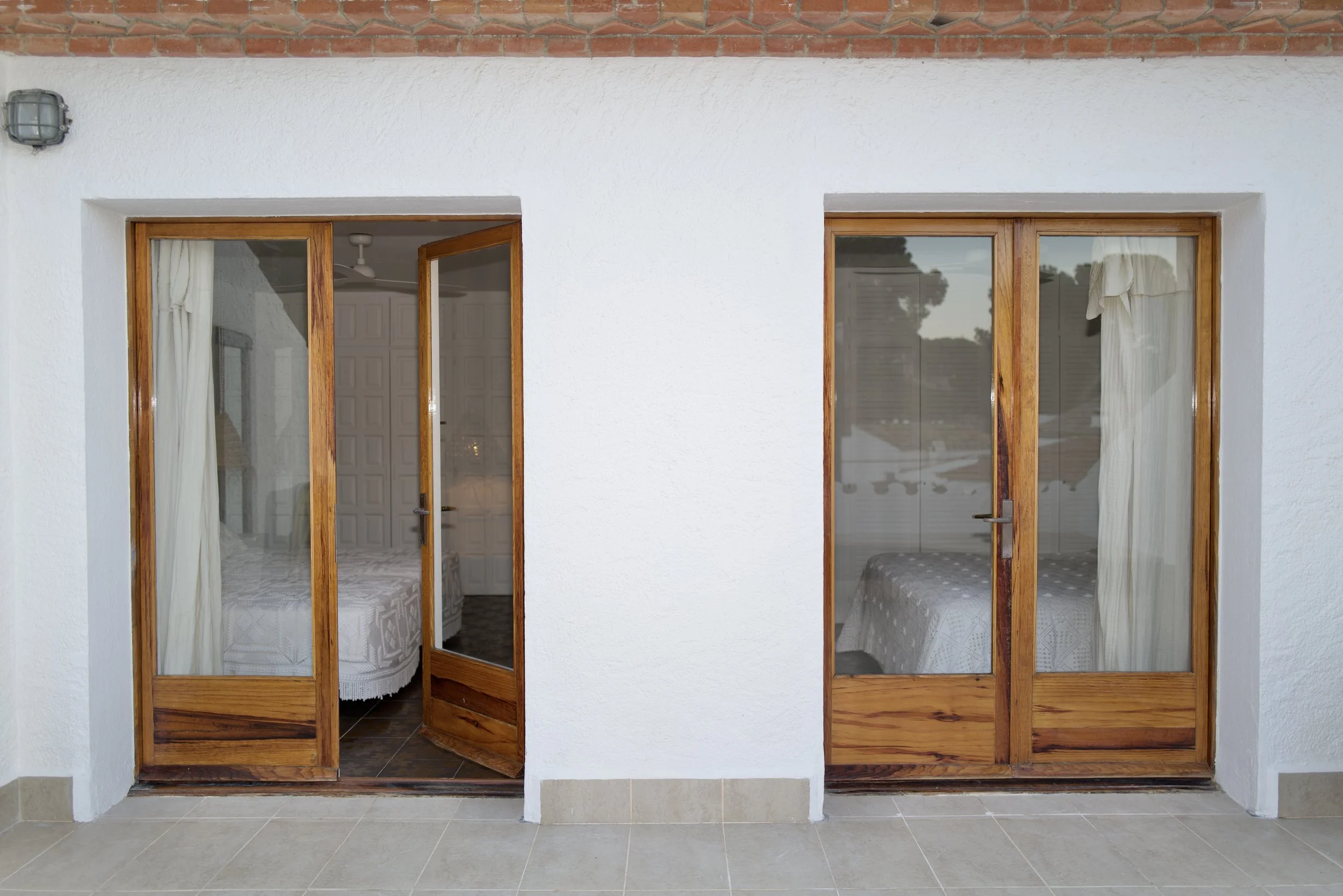 Puertas de vidrio con marco de madera que llevan a habitaciones con cortinas blancas. La pared exterior es blanca con un acabado de textura y una parte de ladrillo expuesto en la parte superior. El suelo es de baldosas de color gris claro.
