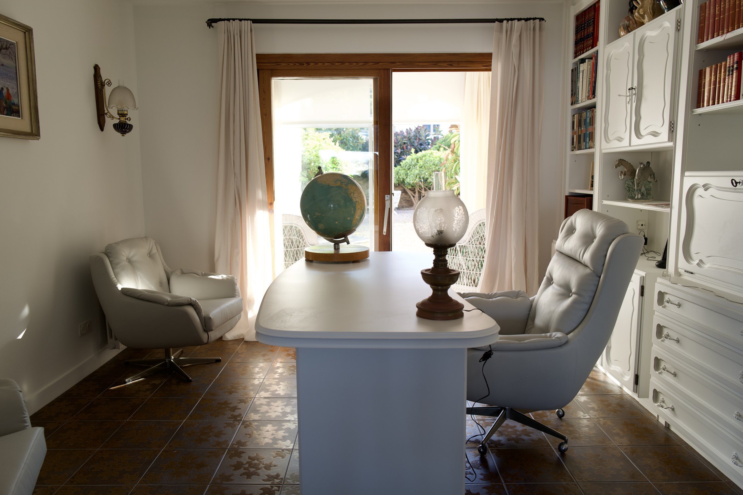Sala de estar con sofá, sillones blancos, mesa de comedor con un globo terráqueo y una lámpara de mesa, con vista a un jardín exterior. Hay estantes blancos con libros y decoraciones.