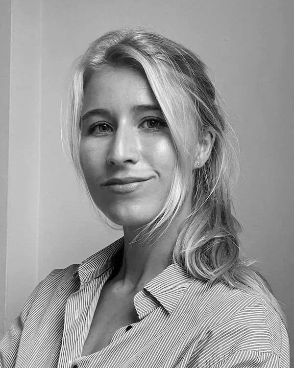 Black and white photo of a woman with blonde hair, wearing a striped collared shirt, smiling slightly, looking directly at the camera.