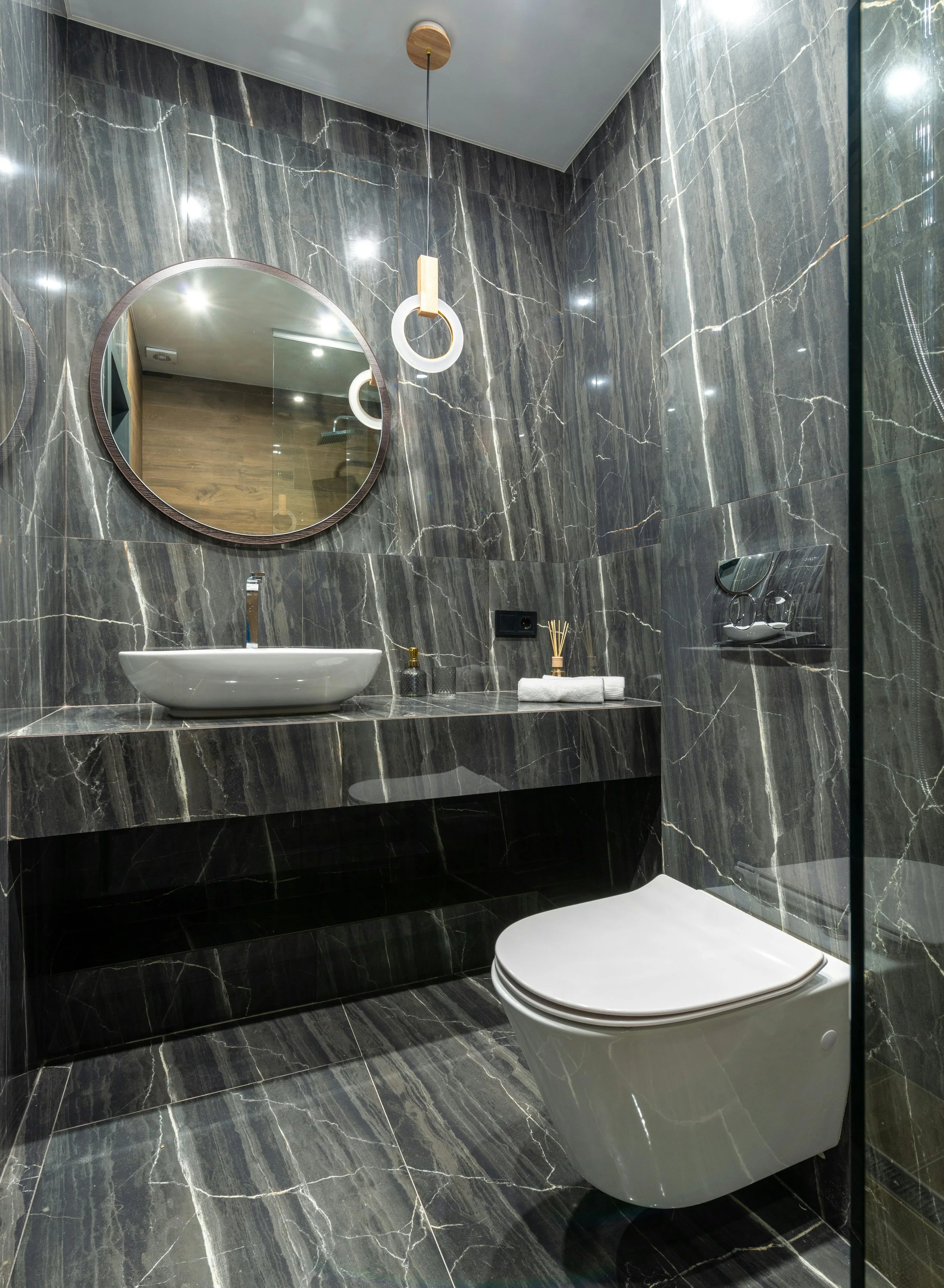 Modern bathroom with black marble walls and floor, a round mirror, white vessel sink, and wall-mounted toilet.