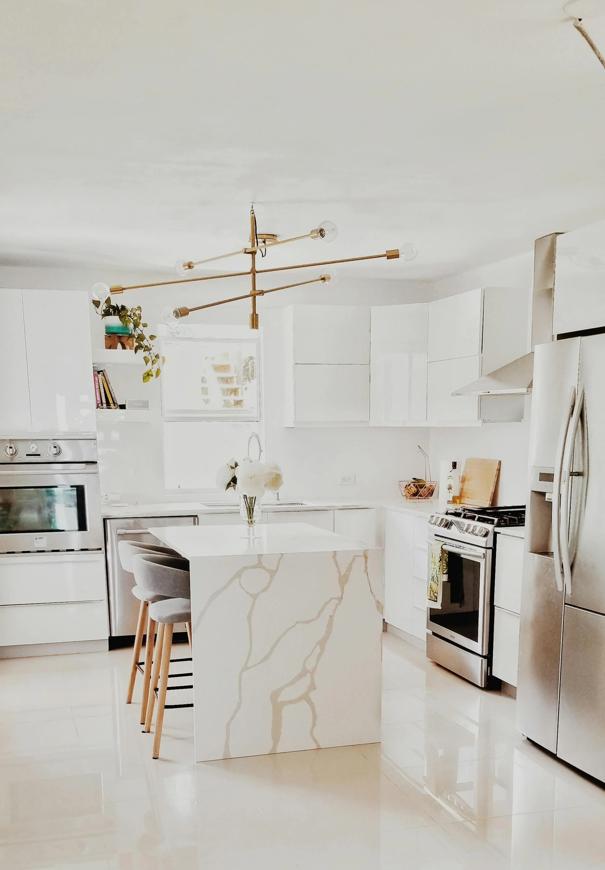 Modern white kitchen with ice blue countertops, stainless steel appliances, and a marble island with gold veining, decorated with a white vase of flowers.