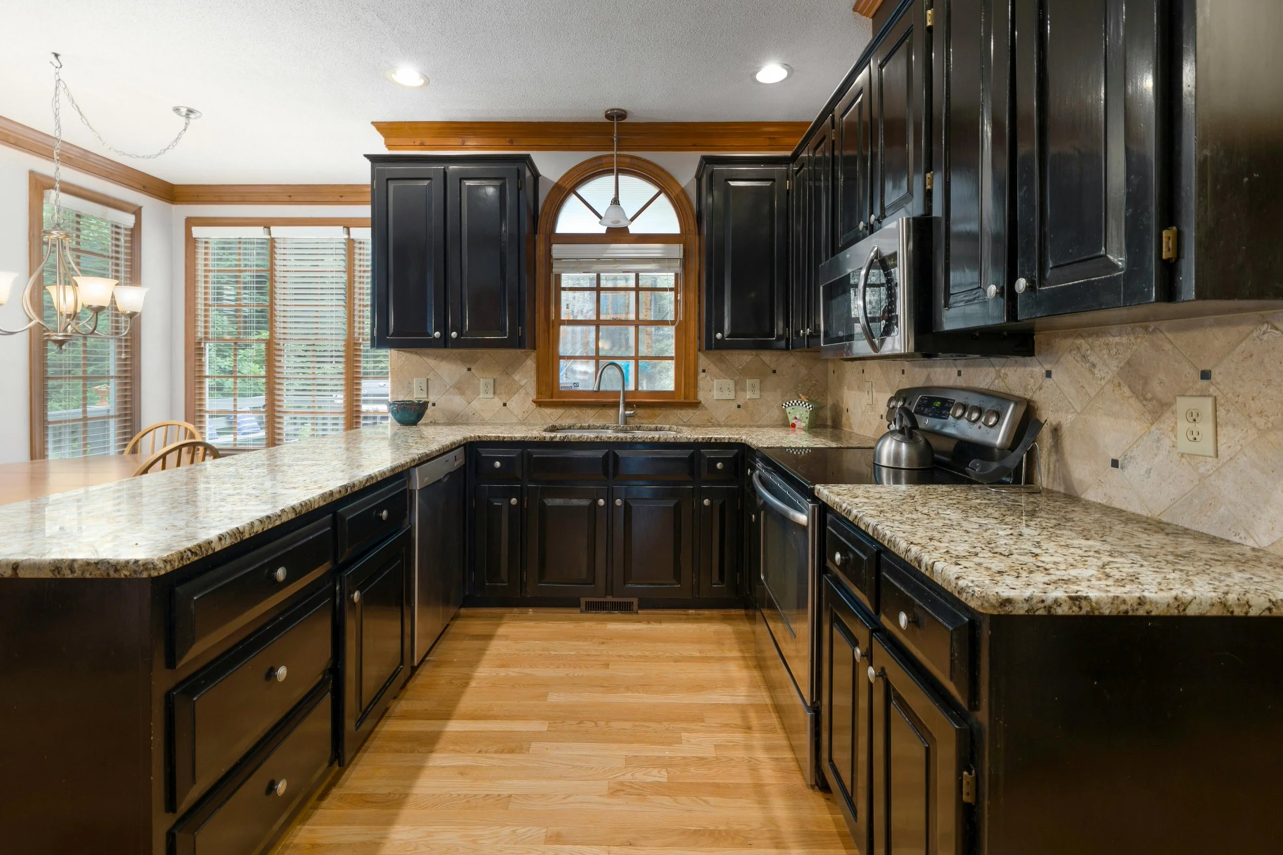 A spacious kitchen with black cabinets, granite countertops, and a window above the sink. There are hardwood floors and a view of large windows and a dining area with a chandelier to the left.