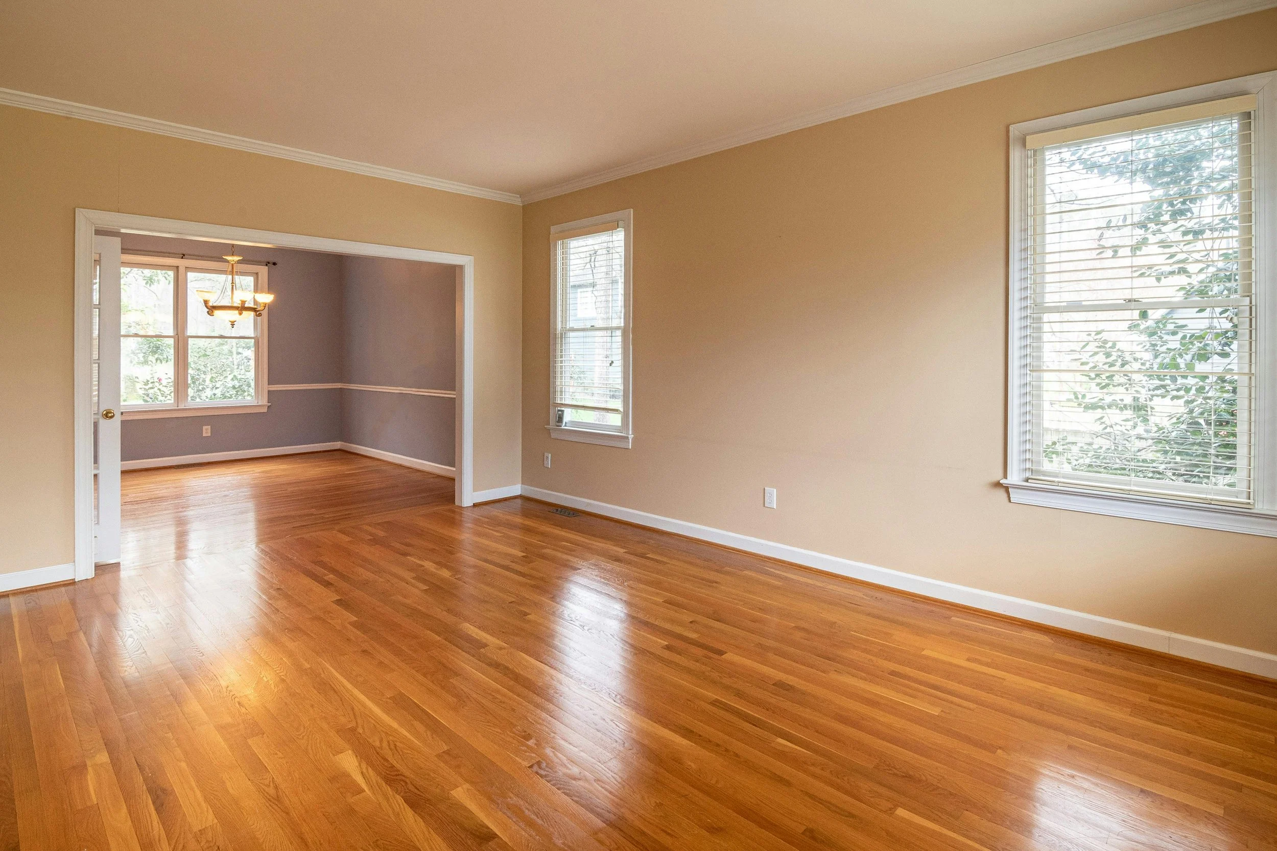 Empty living room with hardwood floors, beige walls, and white trim; two large windows with blinds