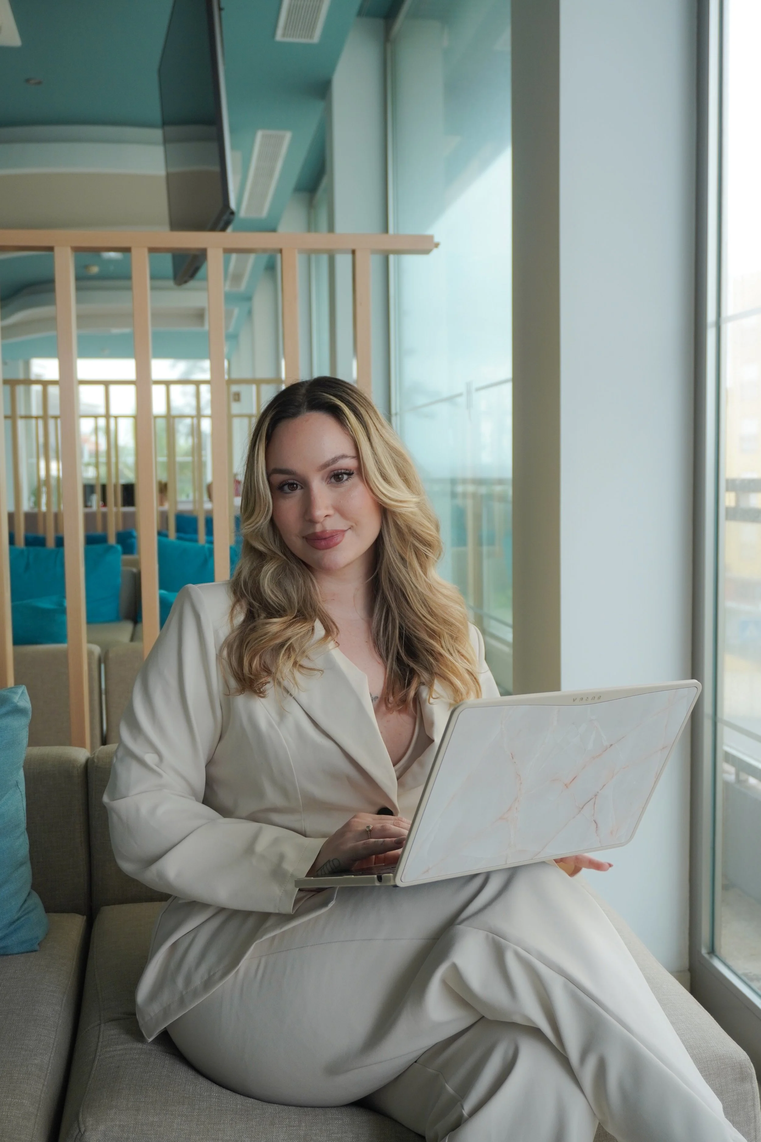Mulher com cabelos loiros e ondulados sentada em um sofá usando roupas brancas, segurando um laptop branco com estampa de mármore, ao lado de janelas de vidro.