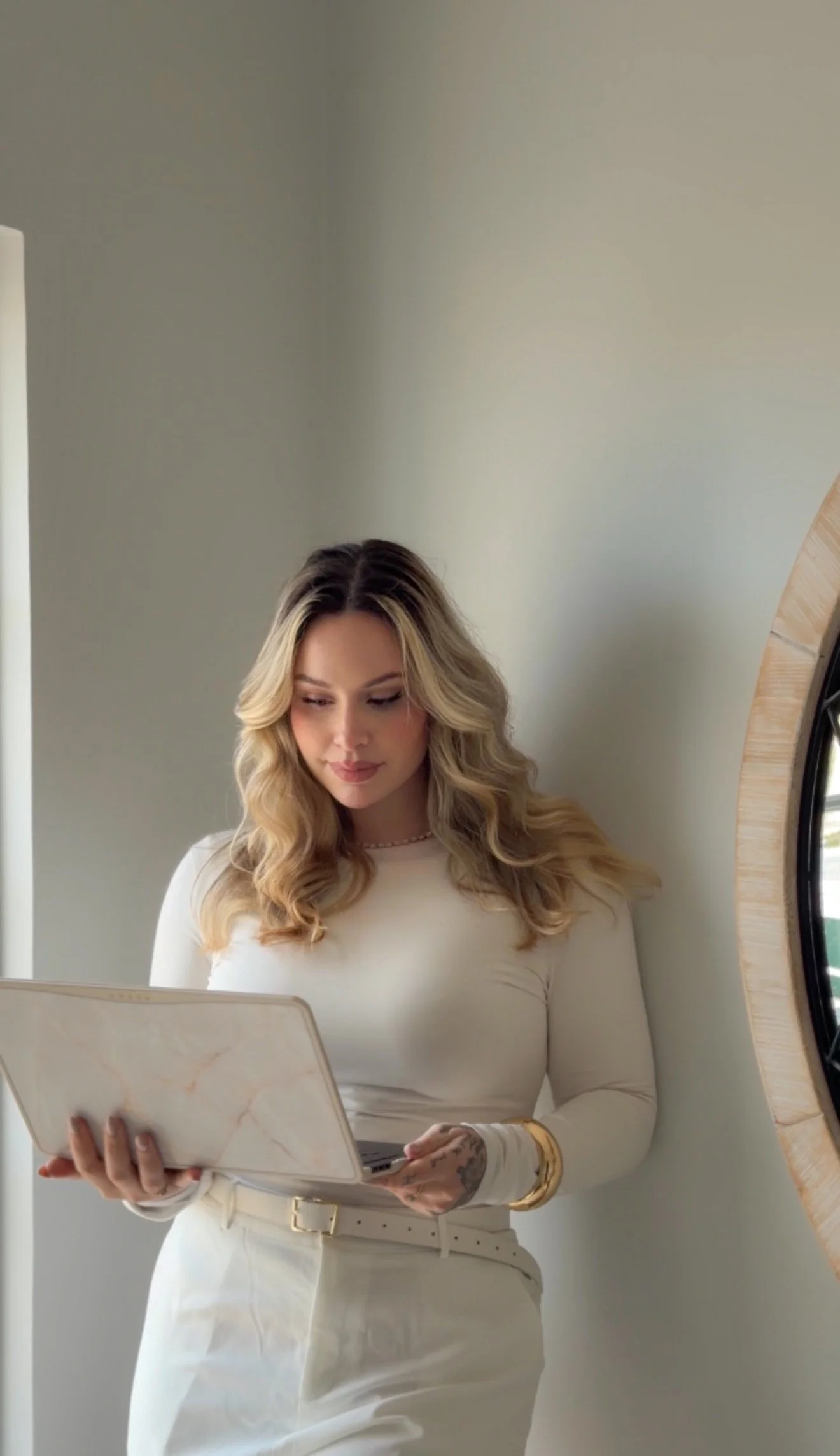 Mulher com cabelo loiro cacheado olhando para um laptop, vestida de branco, com joias douradas, ao lado de uma parede clara e um espelho redondo.