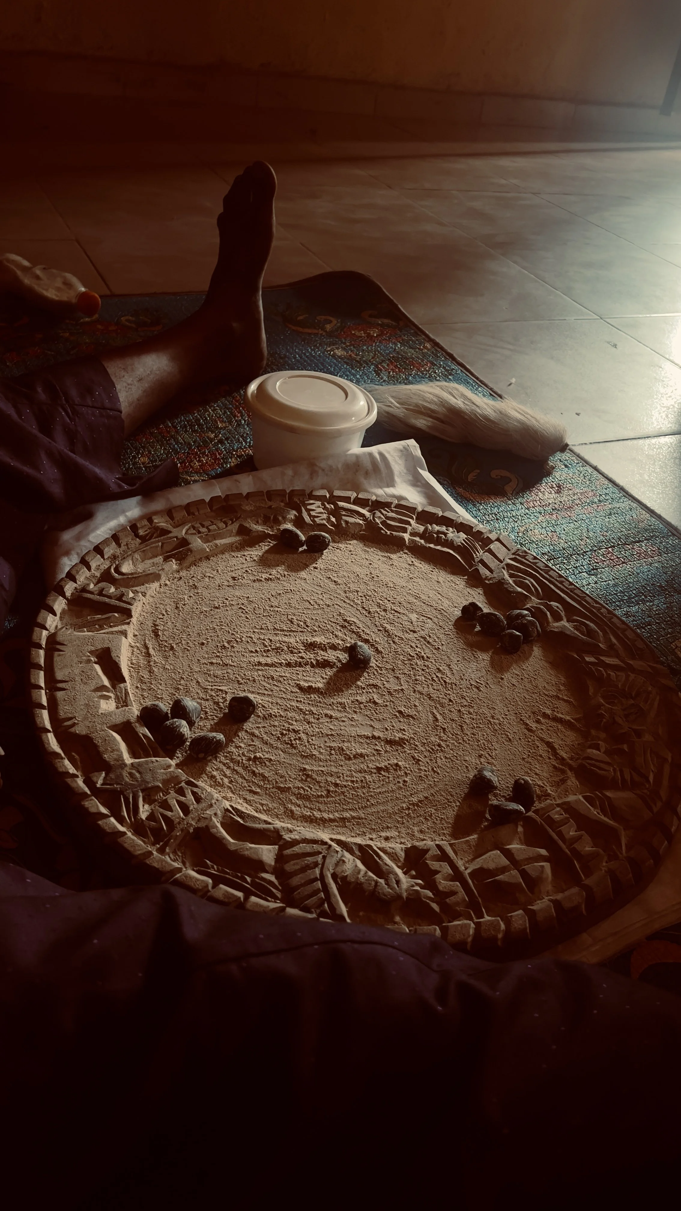 A circular carved wooden tray on a table filled with sand and small stones, with a person's leg and foot, a container, and a white cloth nearby.