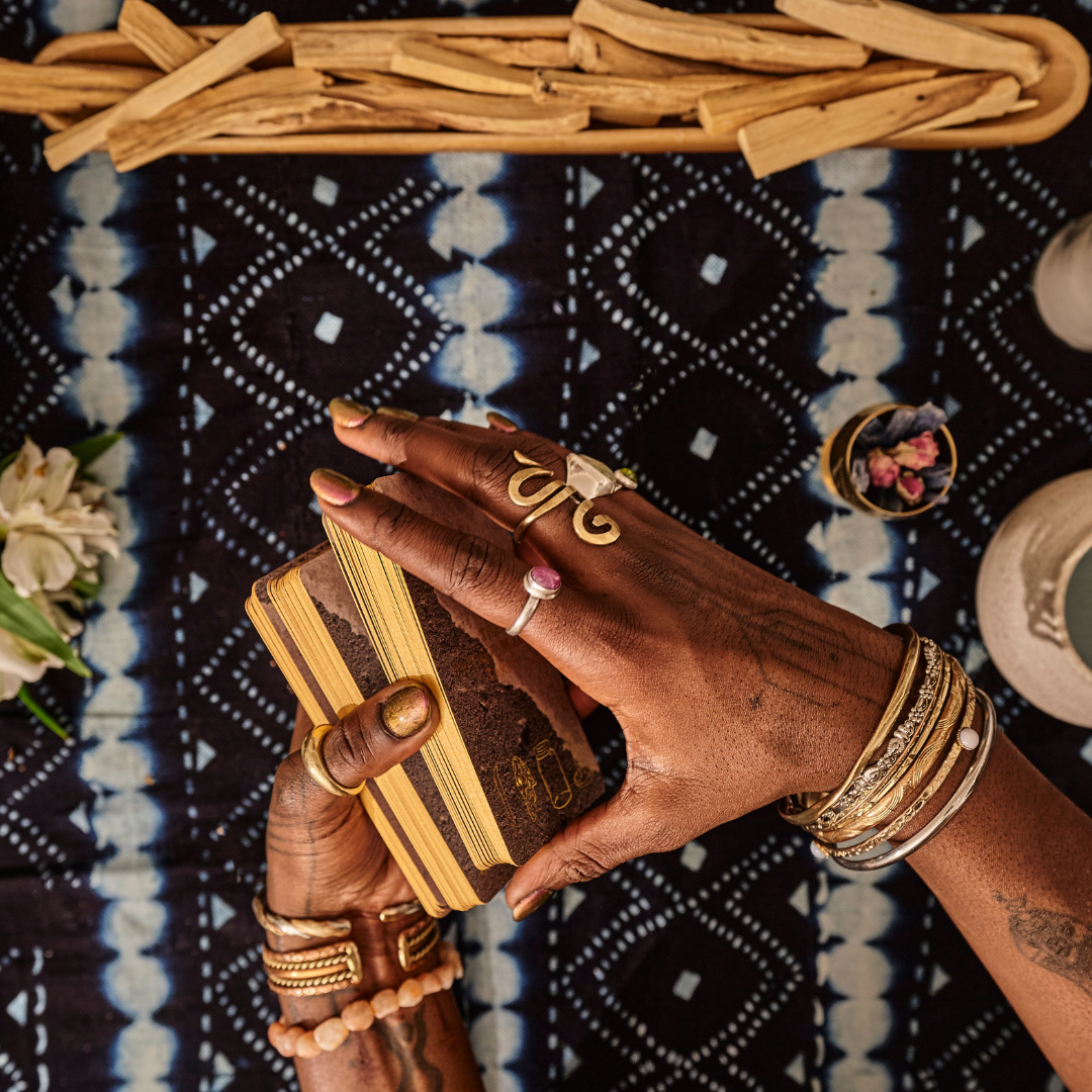 Person with dark skin holding a deck of tarot cards, wearing multiple rings and bracelets, sitting at a table with a patterned cloth, flowers, and decorative items.