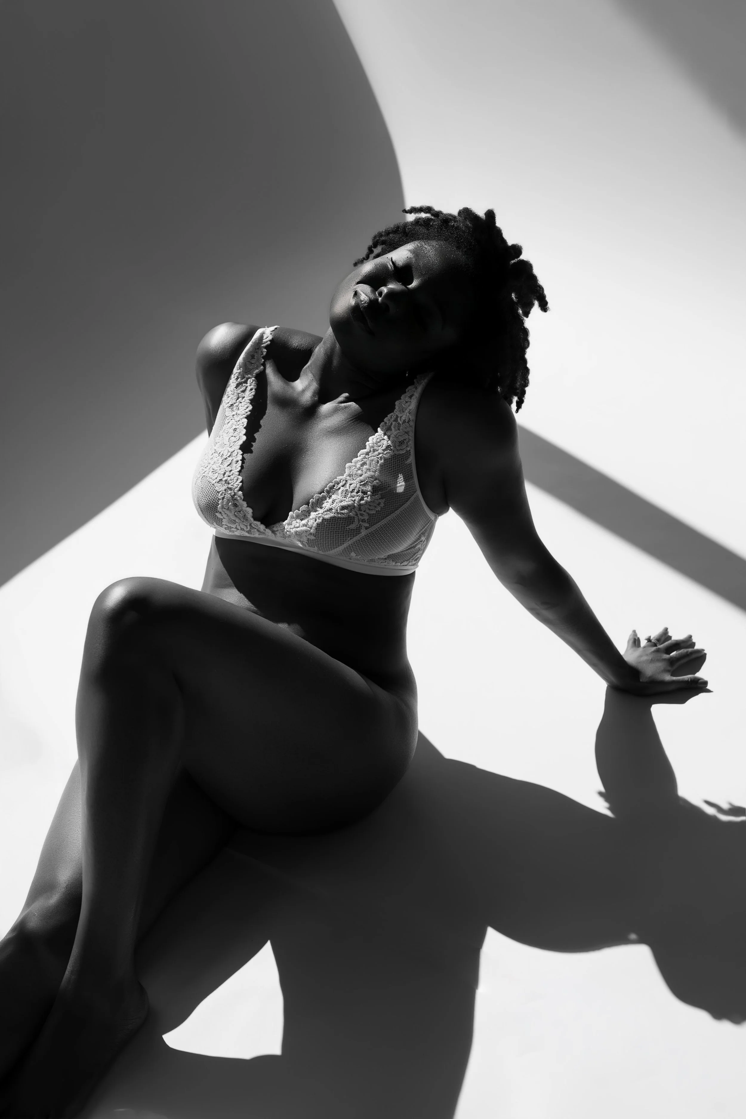 A woman with curly hair in lace lingerie sitting on the floor with shadows cast across her body.